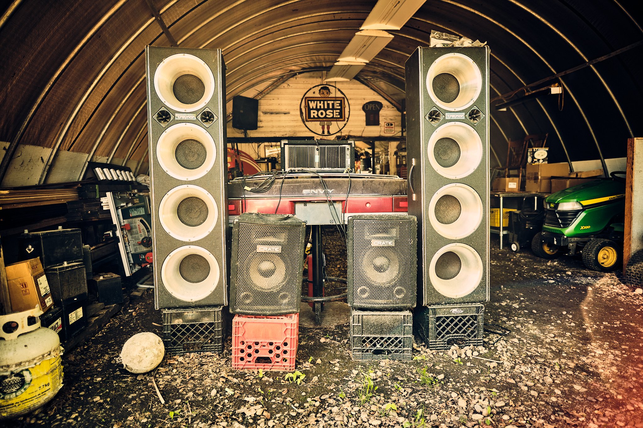 Two large portable speakers set up in a rustic garage or workshop space, with tools and equipment around, and signs including 'White Rose' in the background.