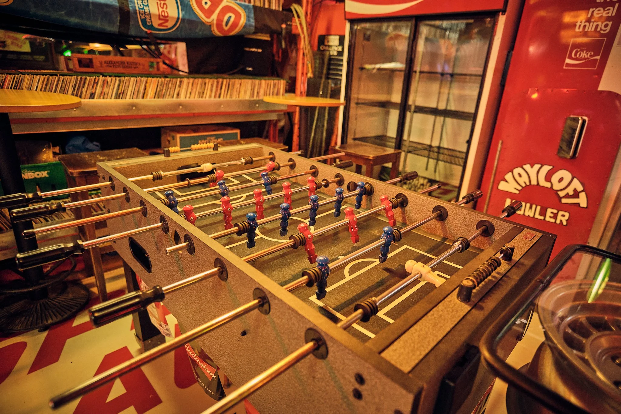 Photo of a foosball table in a room filled with various arcade and vending machines, including a refrigerator and a Coca-Cola vending machine, with stacked shelves and colorful game-related items in the background.