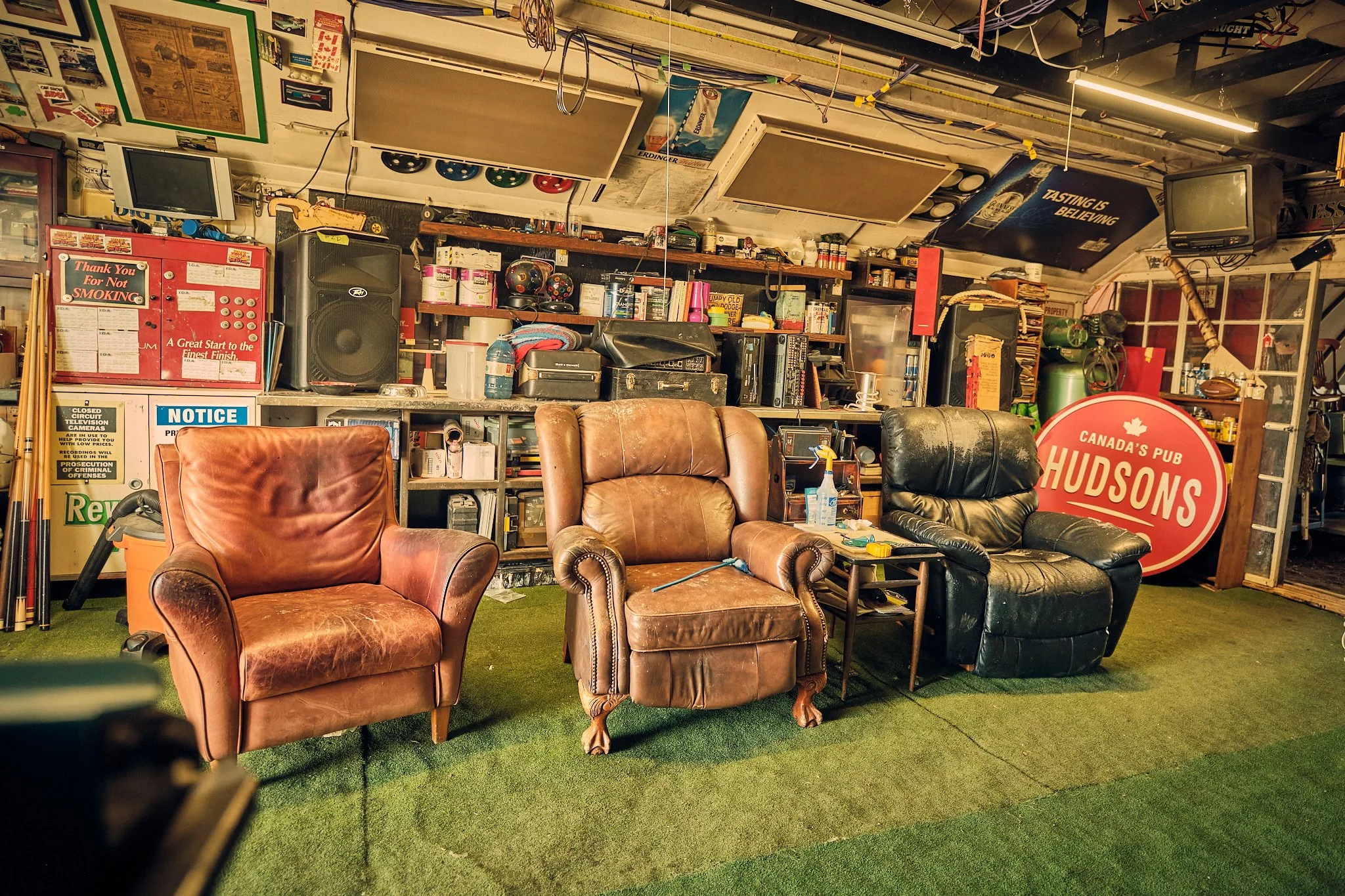 A cluttered garage or workshop with leather armchairs, a black sofa, and various shelves filled with books, cans, and tools. There are multiple old TVs, a sign that says 'Thank You For Not Smoking,' and a red circular sign for 'Hudson's.' The ceiling