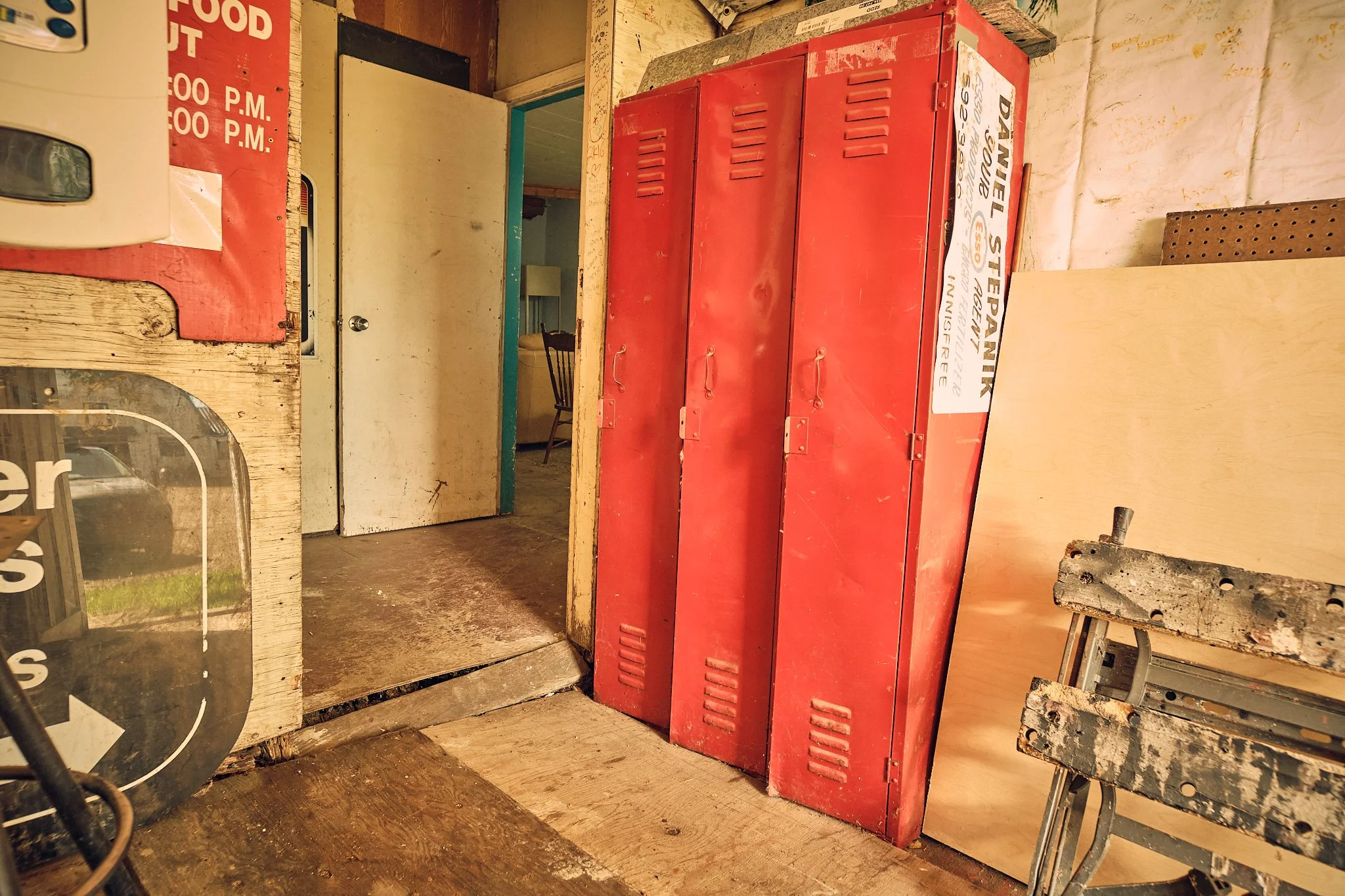 Red metal lockers inside an old rustic room. The room has a wooden floor and walls with visible signs of wear. To the left, part of a sign and a small mirror are visible. Behind the lockers, there is a door and a chair. The room has dated and rustic 