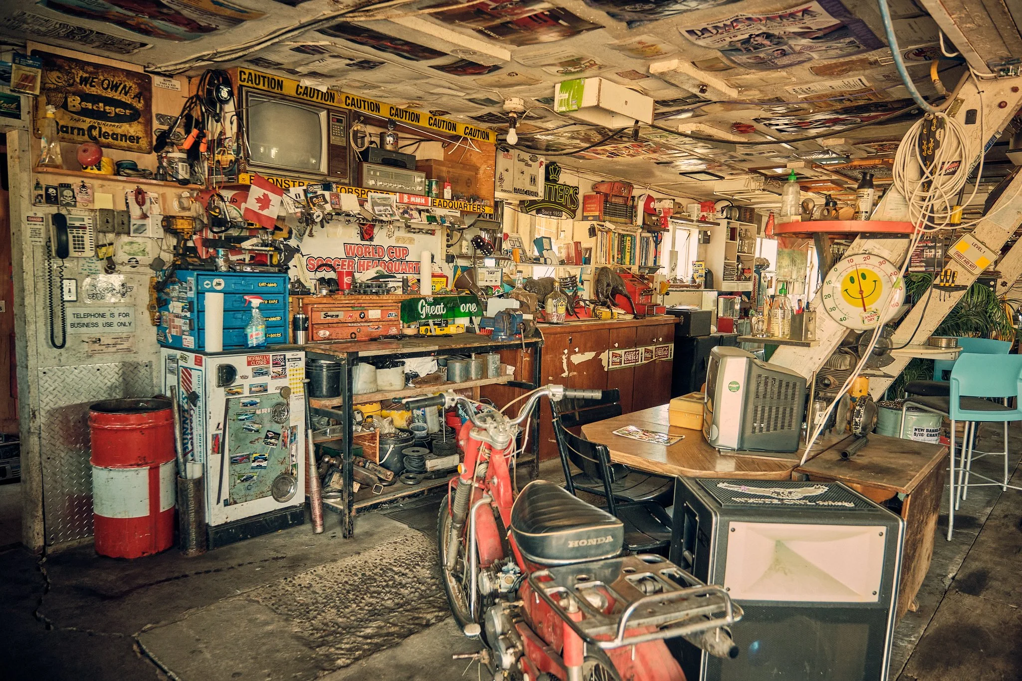 A cluttered workshop or garage with various tools, equipment, and supplies, a red bicycle, and a hoarding of items on shelves and surfaces, with a yellow caution tape border on the wall.