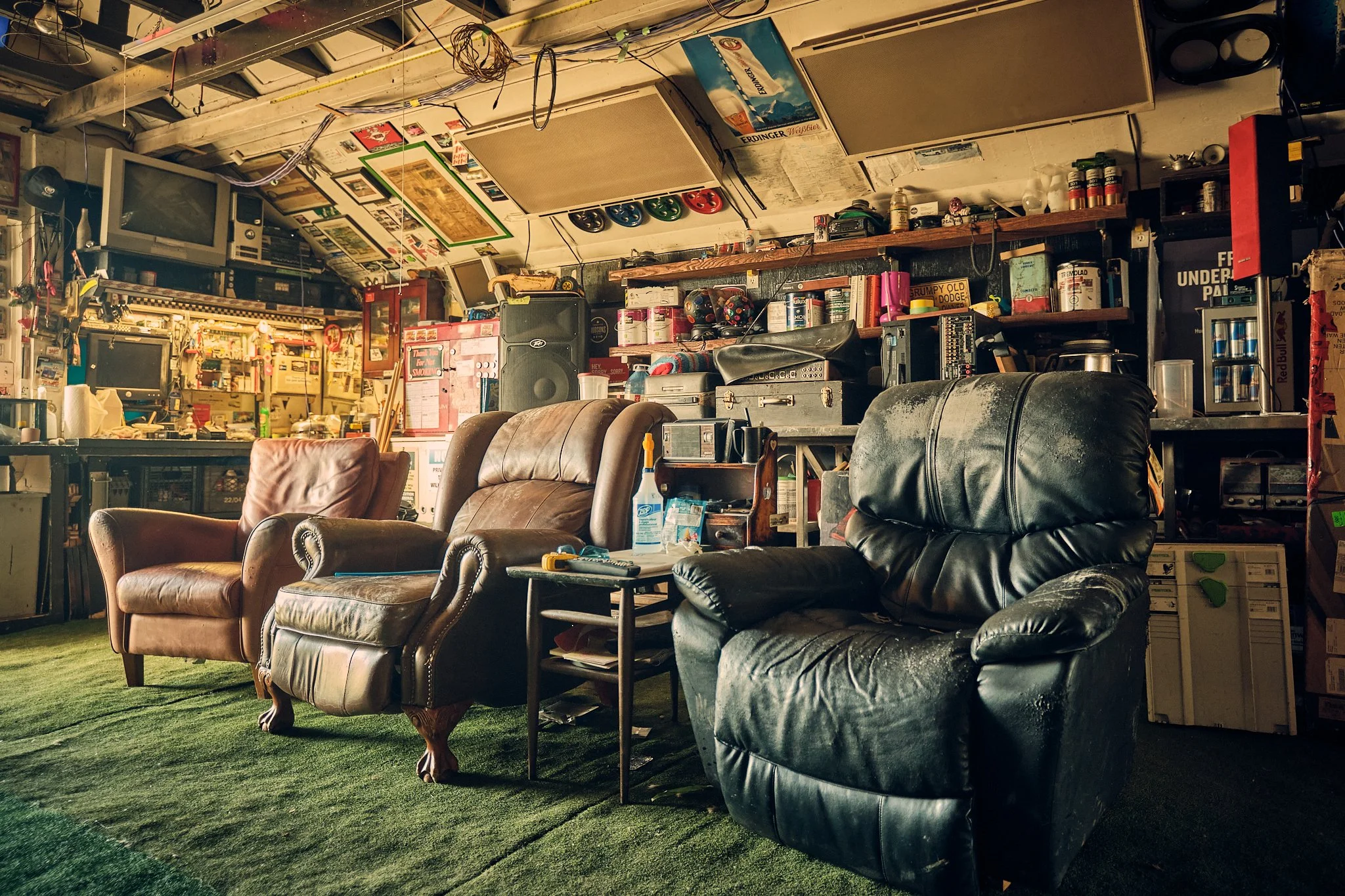 A cluttered room with a mix of furniture, including leather armchairs, a black leather sofa, and a mix of shelves and furniture filled with various items. The ceiling is exposed with wires and ceiling fans, and there are multiple electronic devices a