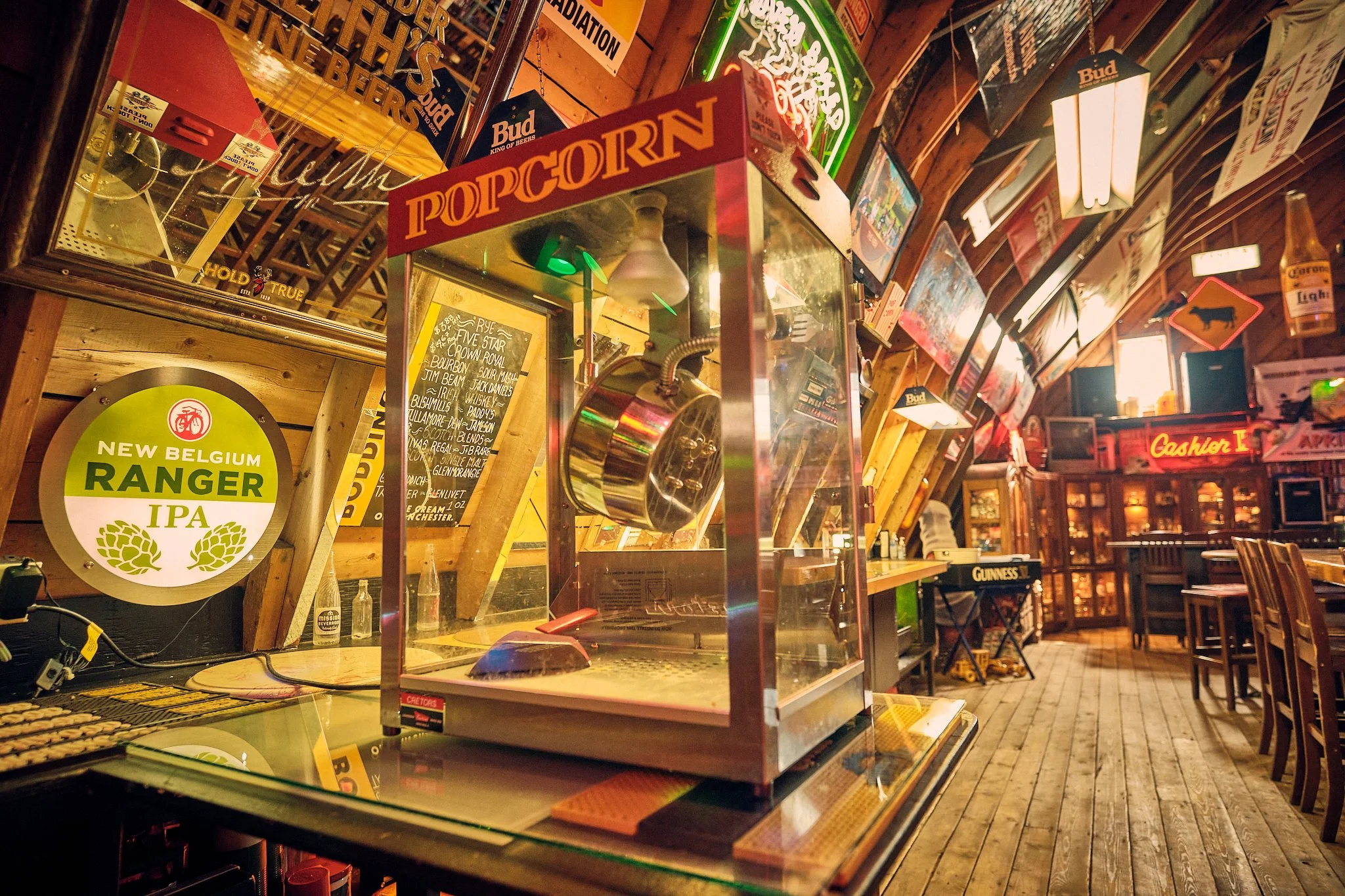 Inside a rustic bar or pub with various neon and wooden signs, including a popcorn machine, beer advertisements, and a chalkboard menu. Wooden walls and tables are visible along with seating.