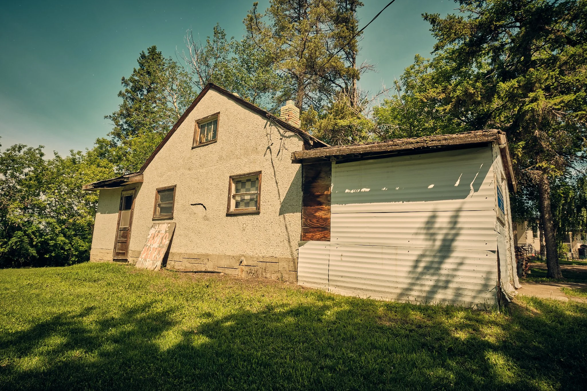 Old house with a gabled roof, multiple windows, and a chimney, surrounded by trees with green leaves, on a sunny day with a clear sky.