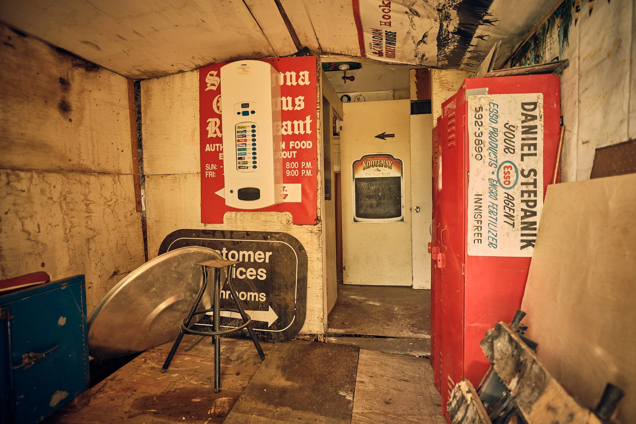 Interior of a rustic eatery with wooden walls, a vintage TV, and various signs, including a red sign and a menu board. There is a small portable heater, some chairs, and a cluttered countertop with a partially burnt piece of paper.