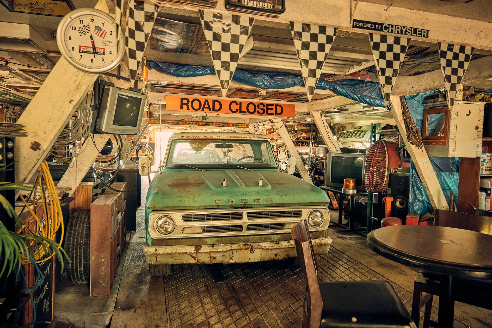 Interior of a garage or workshop with a vintage green pickup truck (Fargo) in the center, surrounded by various tools, equipment, and furniture. A sign hanging from the ceiling reads 'ROAD CLOSED' and another sign says 'Powered by Chrysler.' A clock on the wall shows approximately 5:27. There are checkered pennant banners hanging from the ceiling.