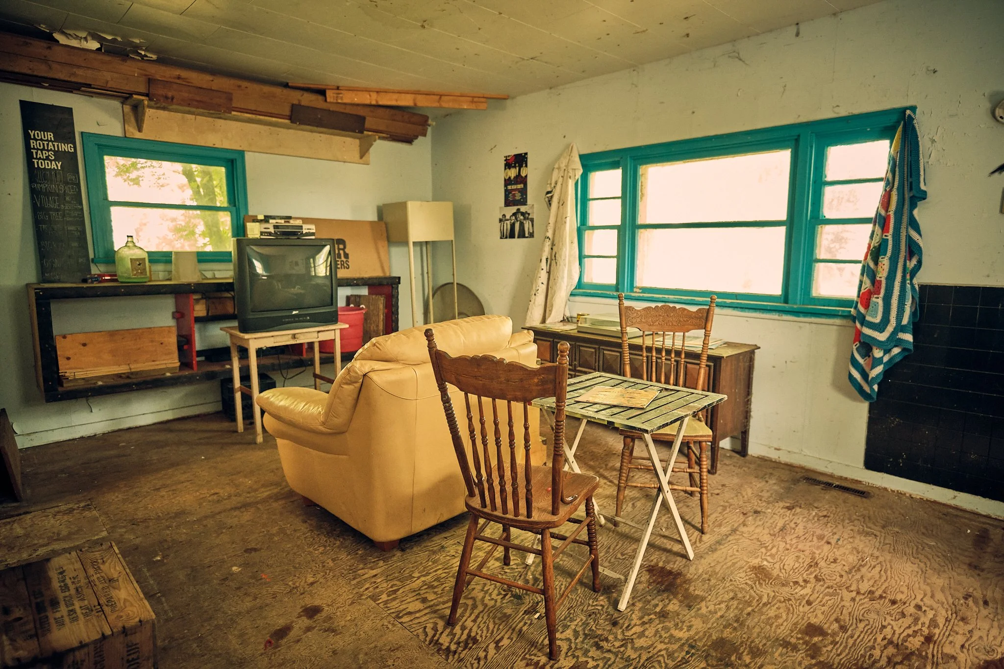 An unfinished room with three large windows with blue frames letting in natural light, a cluttered shelf with a vintage TV, books, and miscellaneous items, a tan sofa, wooden chairs, a small wooden table, and some wall artwork. Part of the ceiling ha