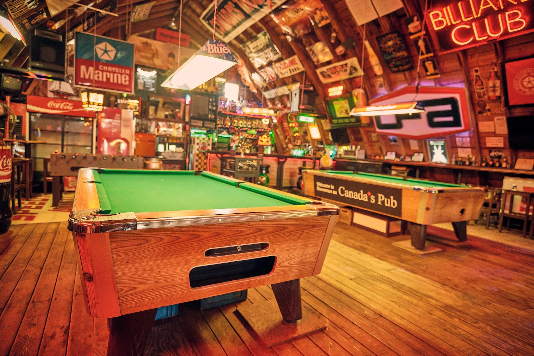 A rustic wood-paneled bar or game room with an American pool table in the foreground, decorated with neon signs, posters, and memorabilia, creating a vintage pub atmosphere.