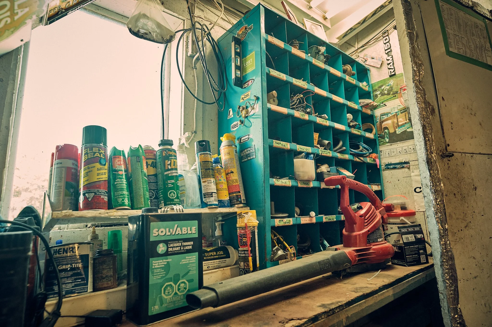 A cluttered workbench with various cans, bottles, and tools, including a red power tool and a blue storage cabinet with small labeled compartments, in a workshop setting.