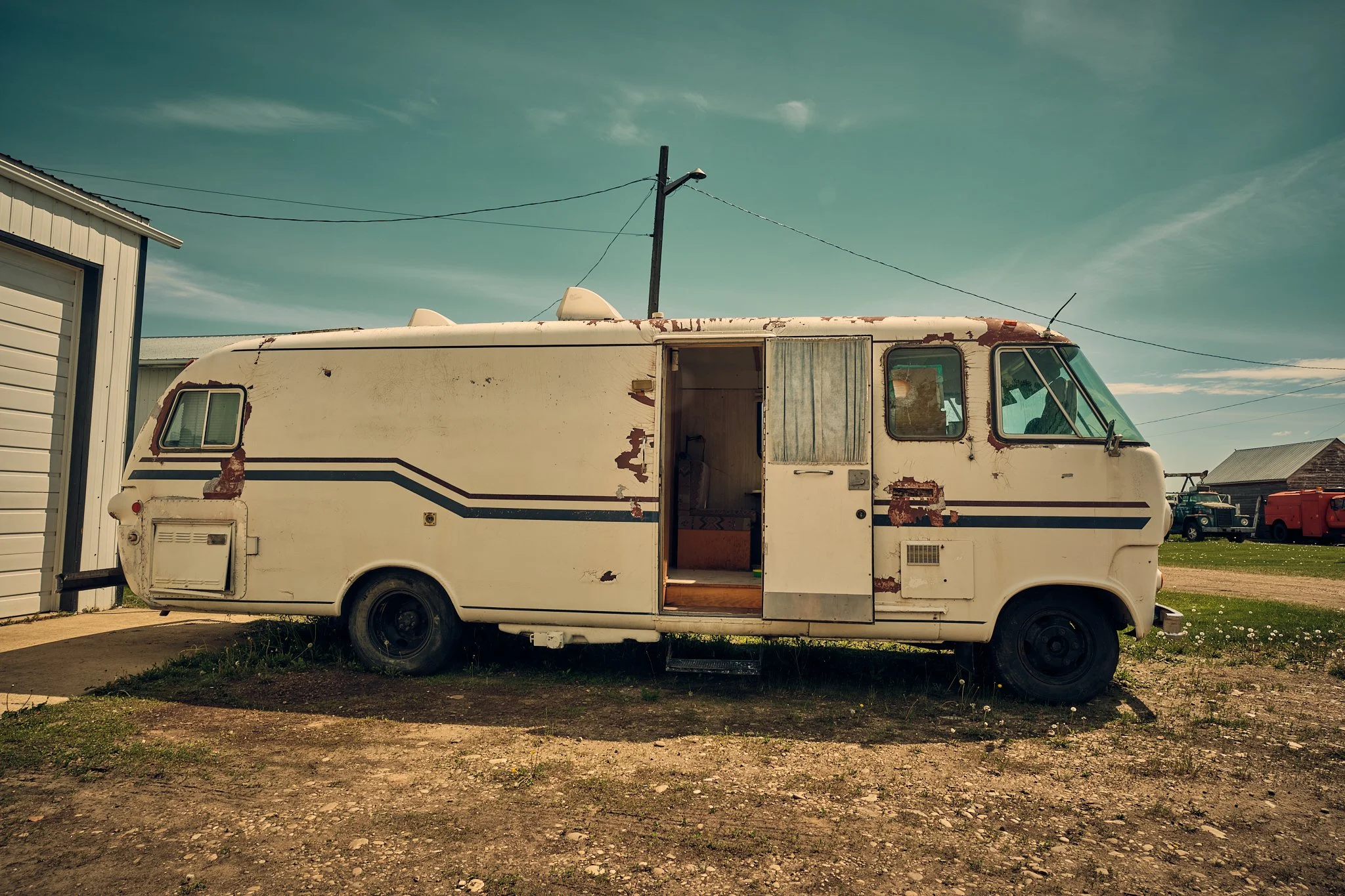 An old, weathered white camper trailer with peeling paint and rusty spots parked on a dirt lot next to a white building, under a partly cloudy sky.
