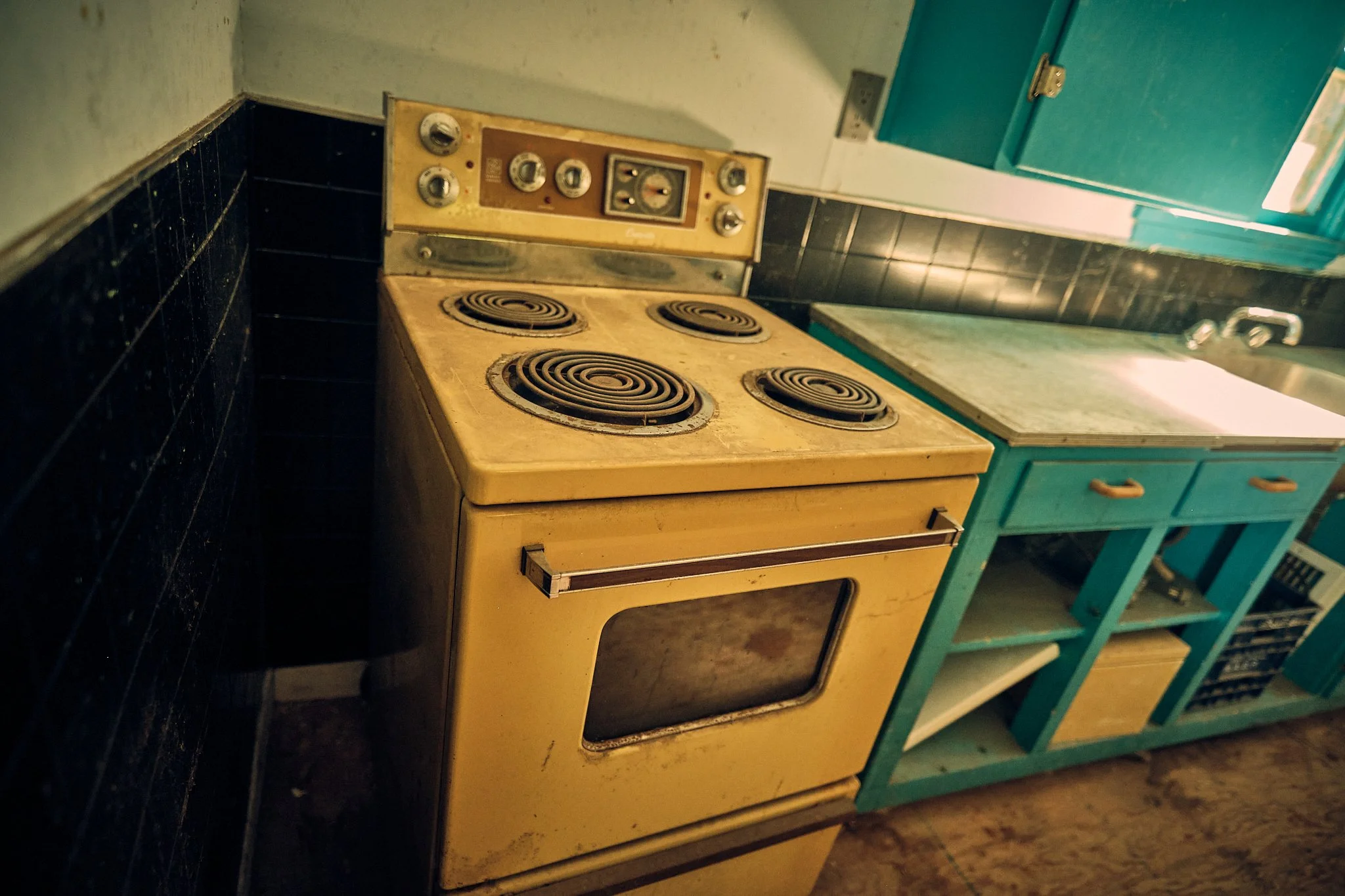 An old, yellow electric stove with four coil burners in a kitchen with black tiled backsplash and turquoise cabinets.