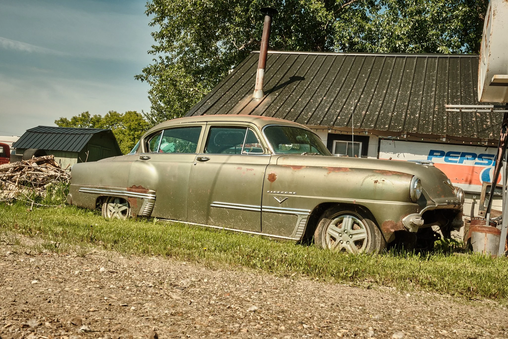 An old, rusty vintage car parked outside a building with a Pepsi sign. The building has a slanted, weathered metal roof and a chimney pipe. Behind the building, there are green trees and a clear sky.