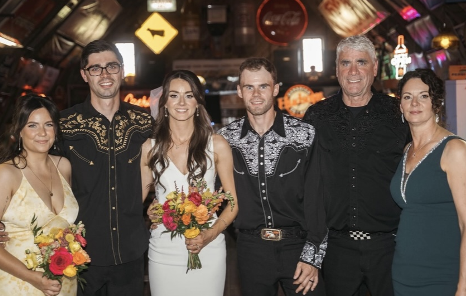 Group of six people at a celebration or wedding, standing indoors with colorful neon signs and vintage decor in the background. They are dressed in formal attire, smiling, with four women holding bouquets of flowers.