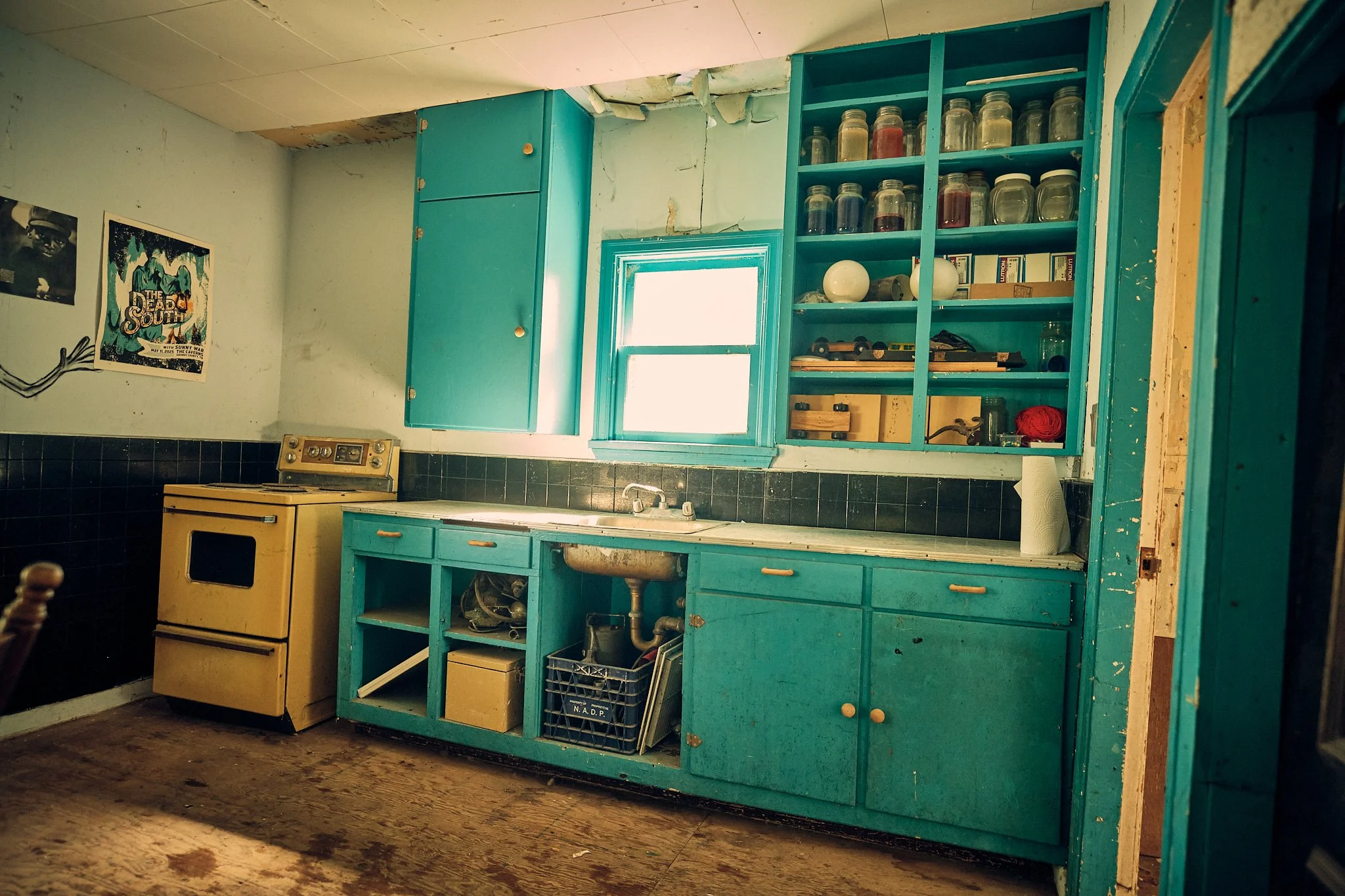 An old kitchen with teal cabinets, black tiled backsplash, and a vintage yellow stove. The cabinets are open, displaying jars and various items. The ceiling and the wall above the cabinets are damaged, with peeling paint and cracks. There is a small 