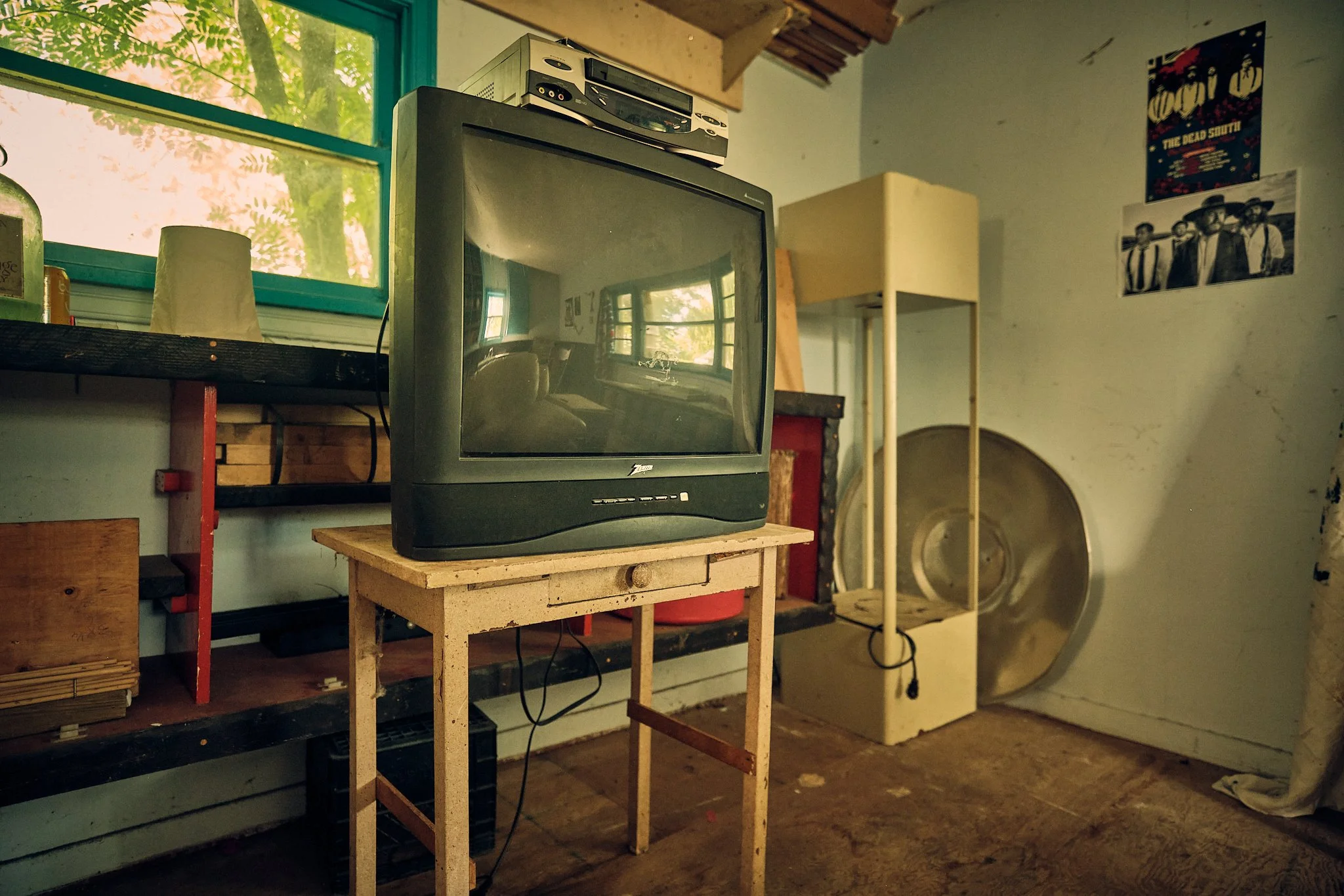 A room with a CRT television on a small wooden table, an old stereo system on top of the TV, and posters on the walls, with a large speaker or drum in the background near a window showing outdoor greenery.