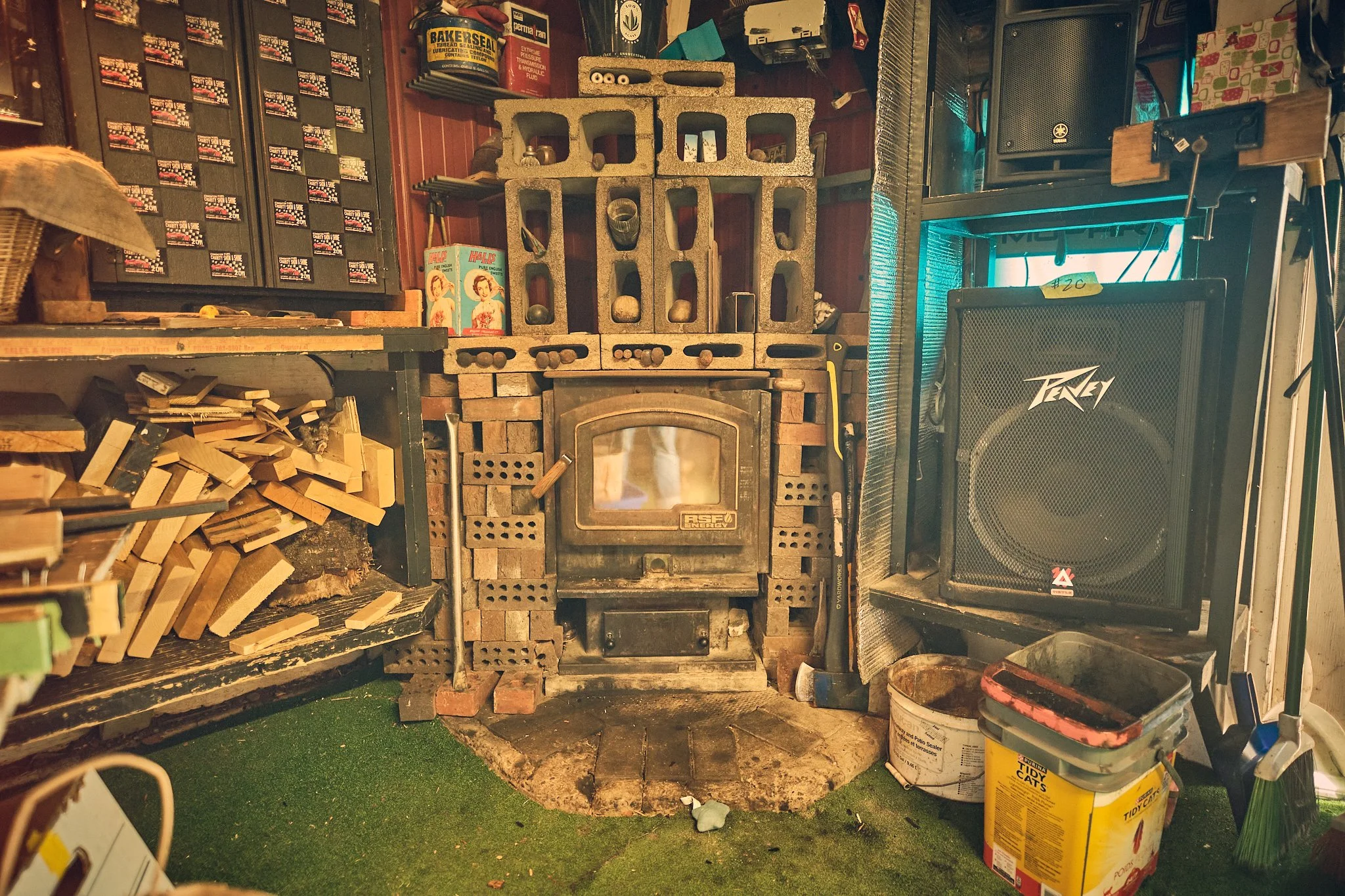 A workshop area with a black wood stove, cinder blocks, and wooden scraps on the floor. Various tools, boxes, and a speaker are visible in the background.