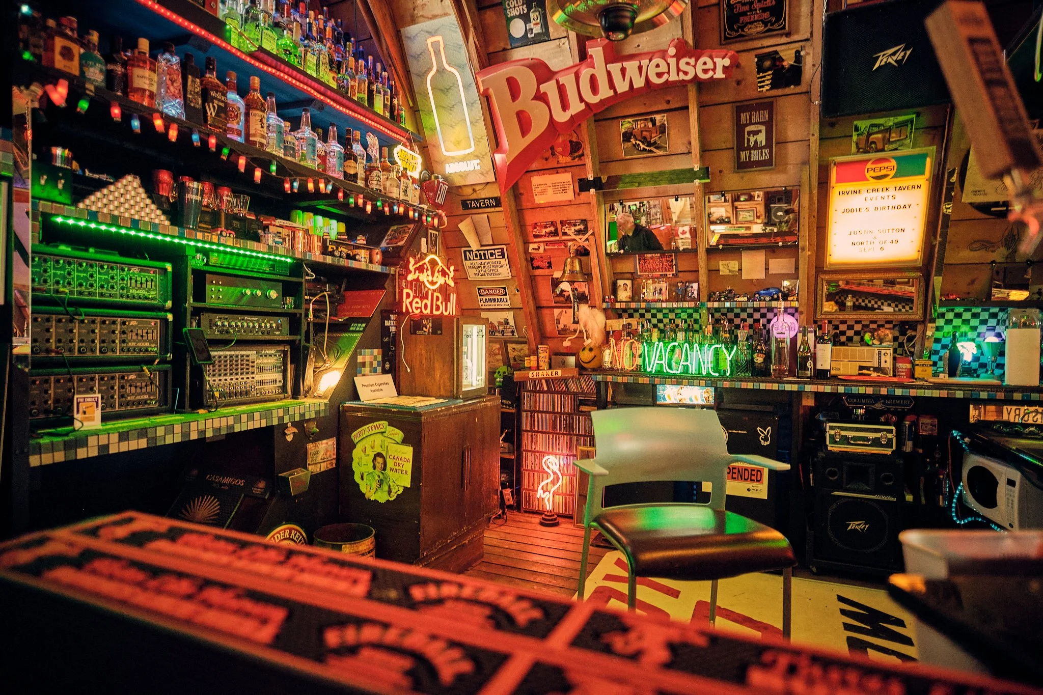 Interior of a bar decorated with neon signs and bottles of alcohol on shelves, with various posters and signs on the wooden walls, including a large Budweiser sign and a neon 'VACANCY' sign.