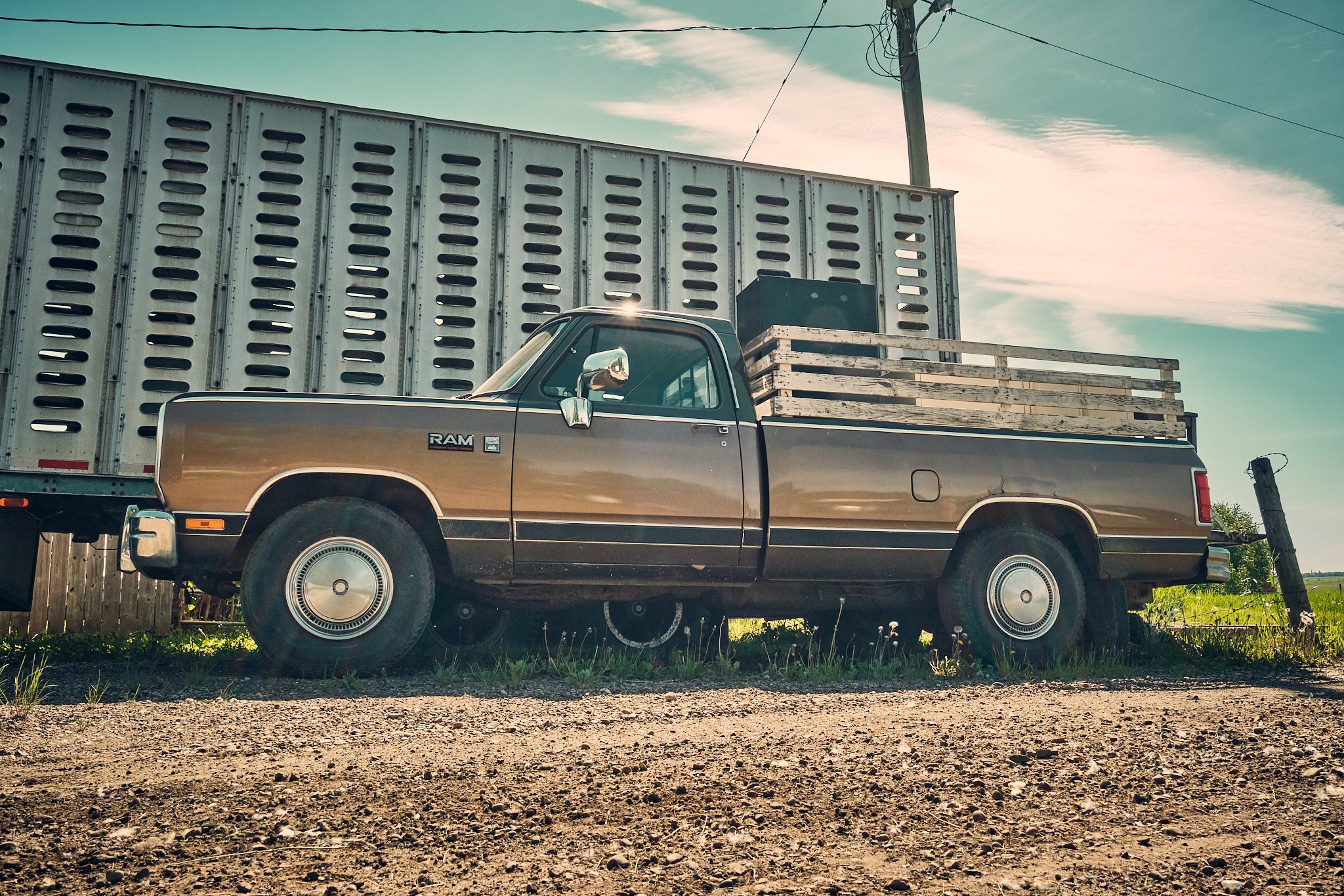 A vintage brown pickup truck with a wooden flatbed parked on a gravel lot, with a large metal lipper behind it and a utility pole in the background against a partly cloudy sky.