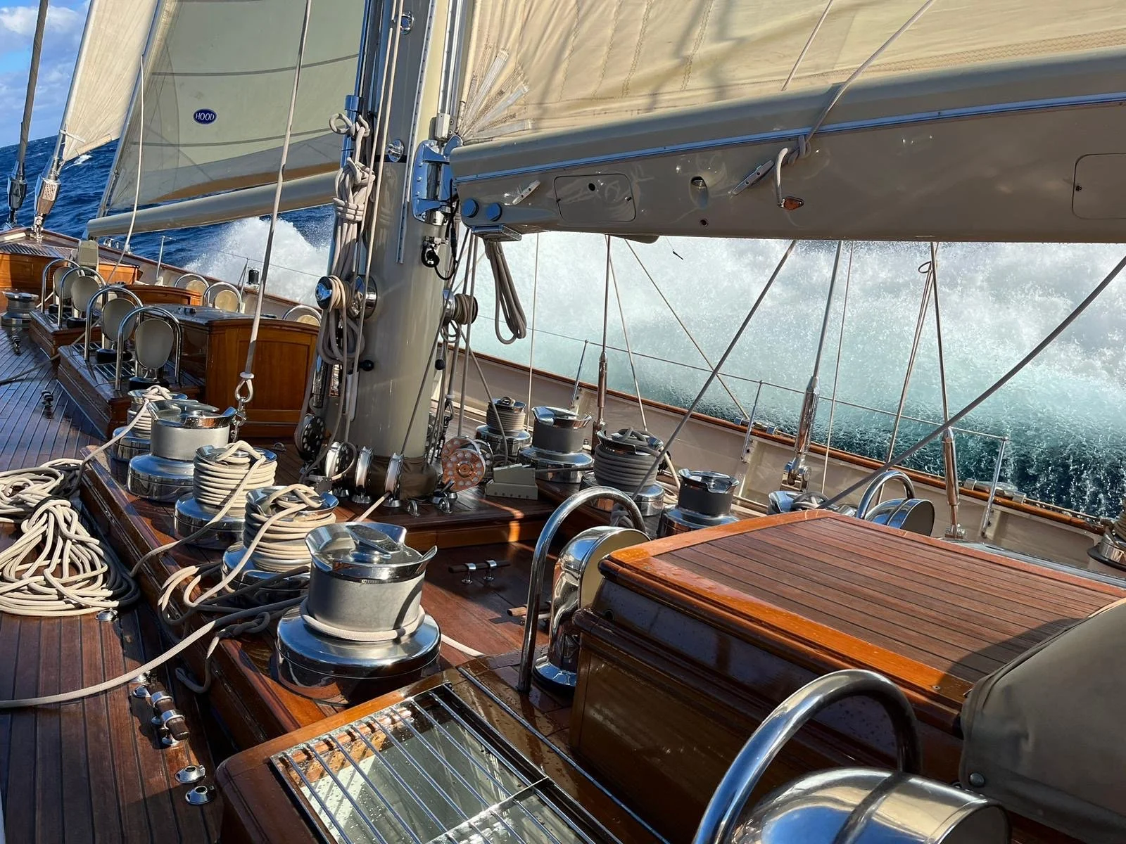 View of a sailboat deck with rigging, winches, and a large sail, with ocean waves and spray in the background.