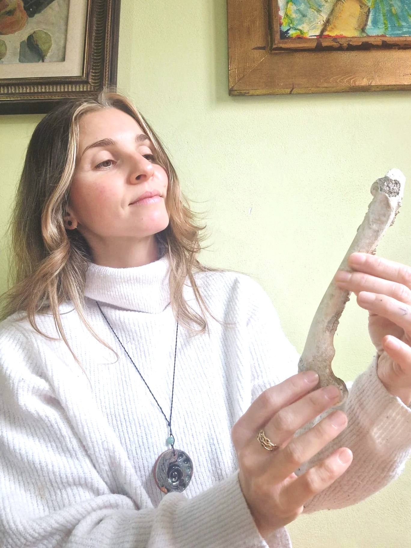 A woman in a white turtleneck sweater holding a large, light-colored bone with a rough surface. She is standing against a light green wall with framed artwork hanging above her.