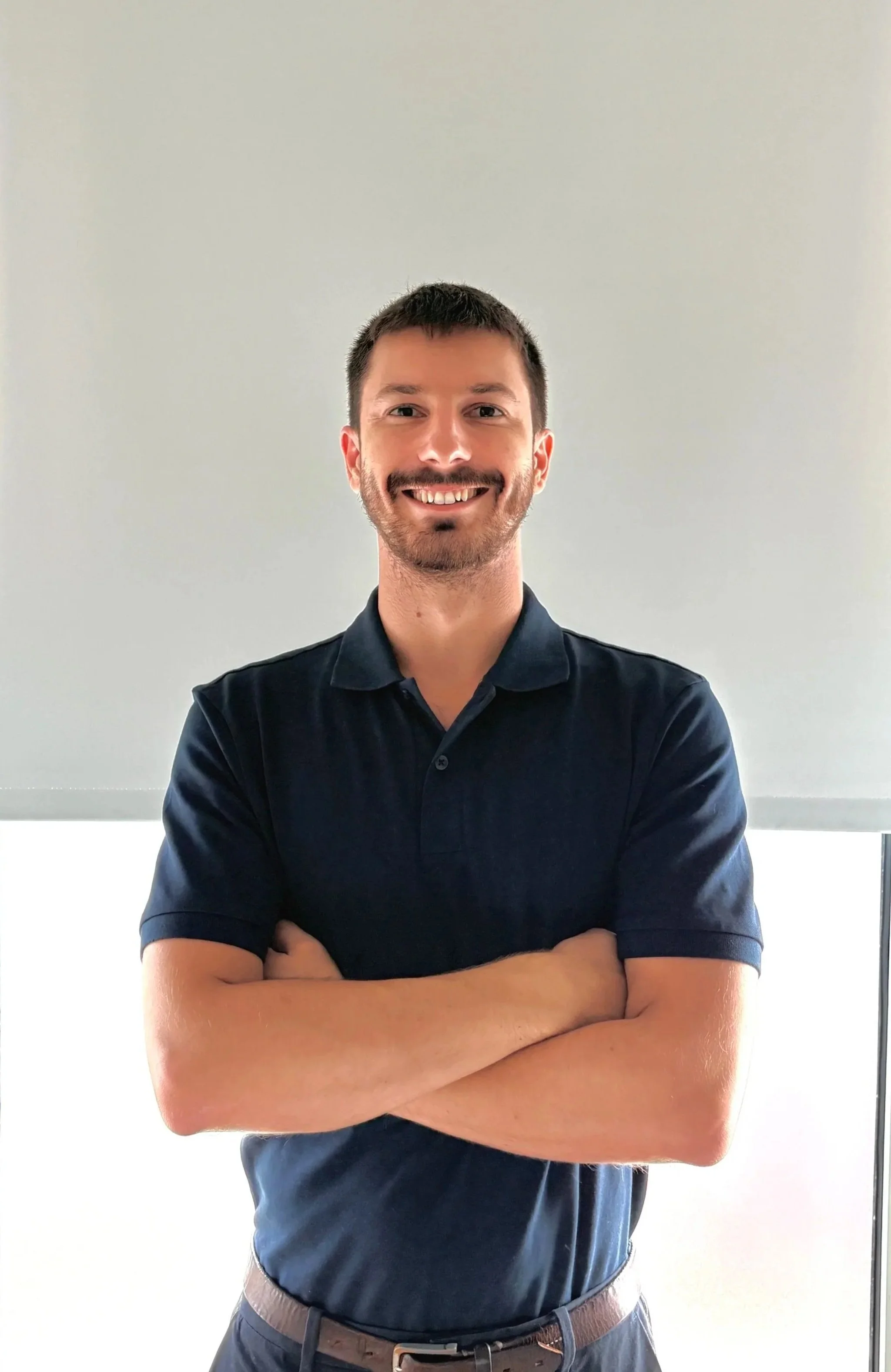 A man smiling with crossed arms, wearing a dark polo shirt and belt, standing in front of a plain light-colored wall.