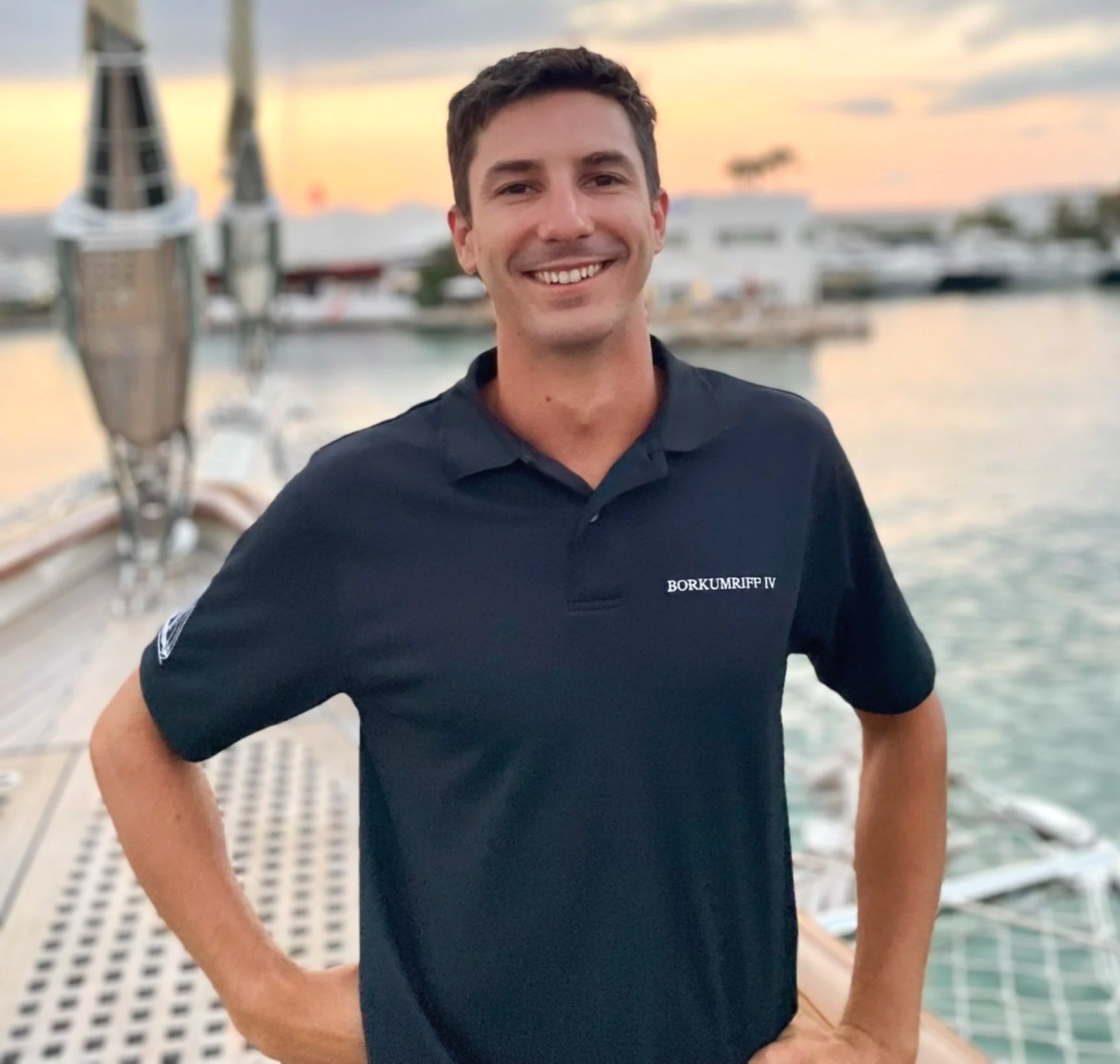 A smiling man in a navy blue polo shirt standing on a boat during sunset, with a marina and boats in the background.