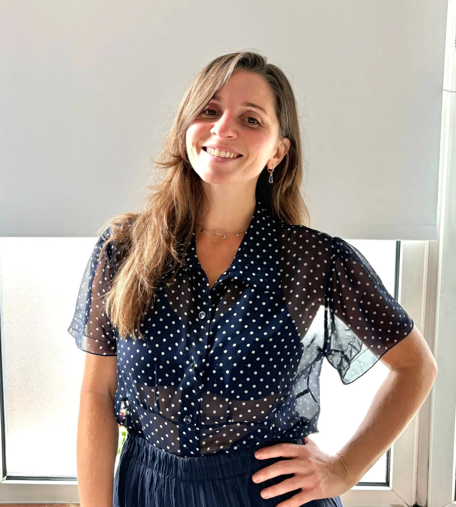 A smiling woman with long, light brown hair, wearing a navy blue polka dot blouse with sheer sleeves, standing indoors near a window with blinds, with her hand resting on her hip.