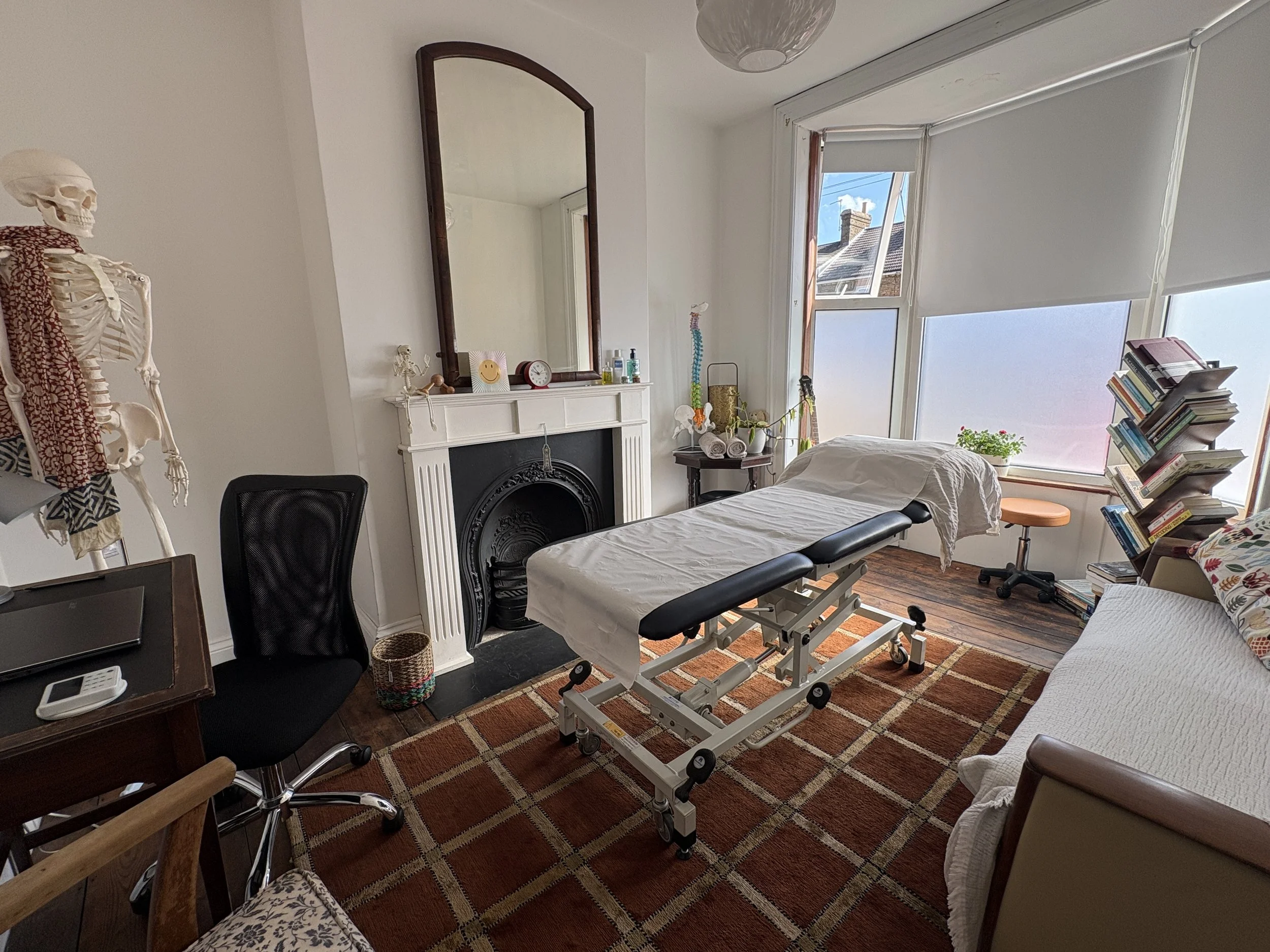 A room with a treatment table, a bookshelf with books, a skeleton model, a large mirror above a fireplace, and large windows with blinds letting in natural light.