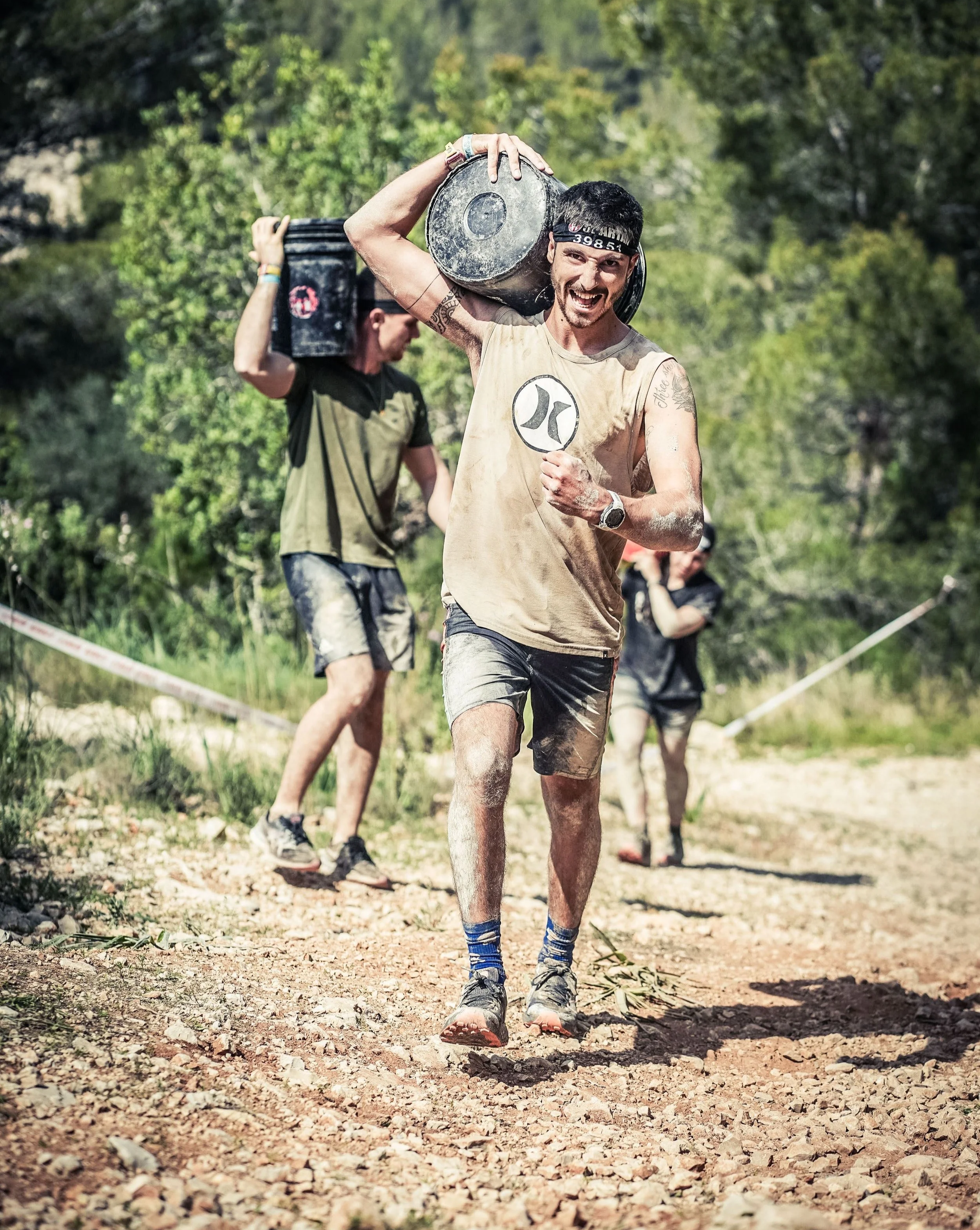 Man participating in an outdoor obstacle course, carrying a large weight on his shoulder, with trees and other participants in the background.