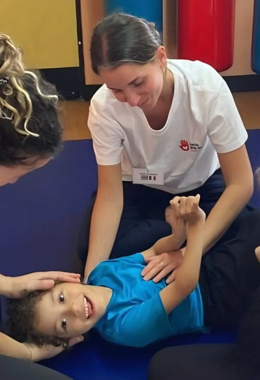 A young girl lying on the floor, smiling, while an adult woman and another person assist her in a first aid or CPR training. The girl is wearing a blue shirt, and the woman is in a white shirt with a red and black logo, wearing a name tag. The scene is indoors with colorful cabinets in the background.