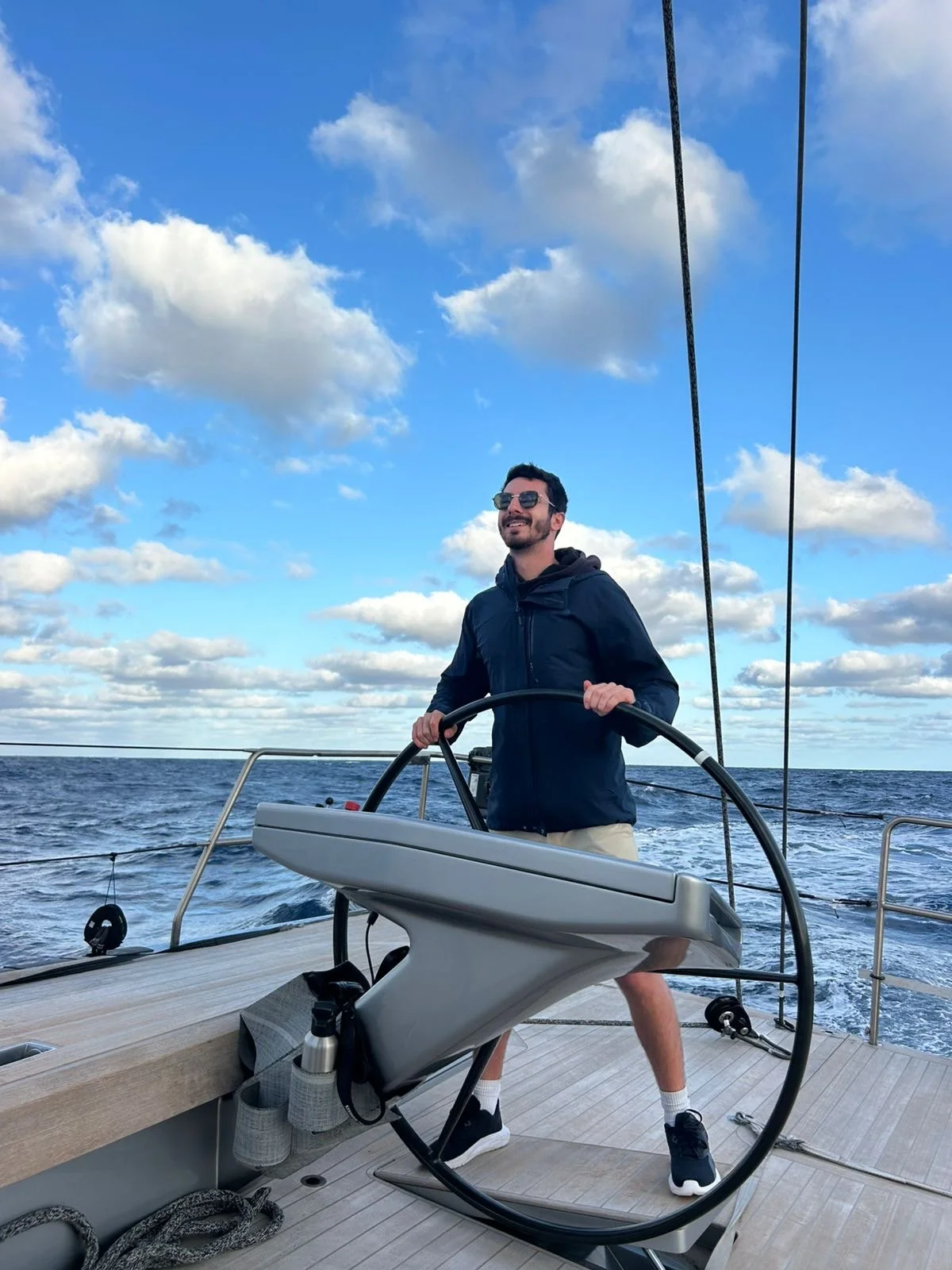 A man steering a sailboat on the ocean, smiling, wearing sunglasses, a dark jacket, and shorts, with a partly cloudy sky in the background.