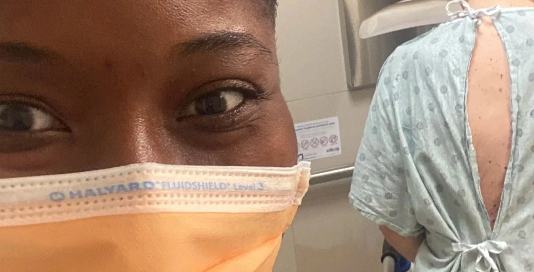 A person wearing an orange disposable face mask and a healthcare worker in scrubs is seen in the background, standing in what appears to be a medical setting.