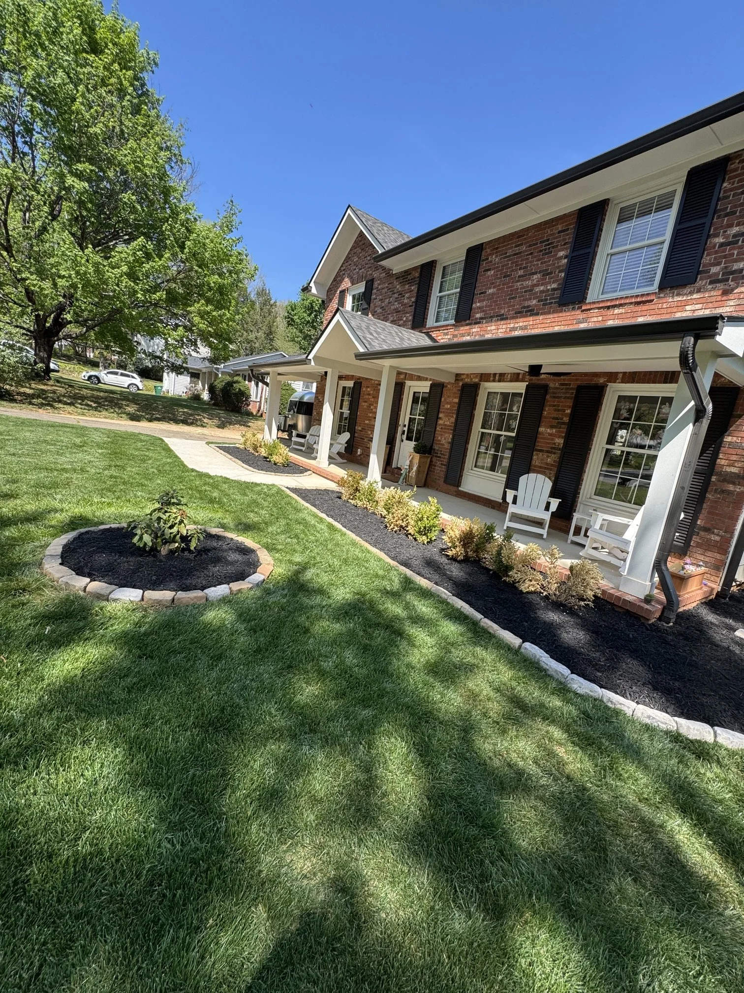 Front yard with green lawn, small garden bed with mulch and plants, brick house with black shutters, white porch chairs, patio, and a large tree near the driveway with a white car parked in the background under a bright blue sky.