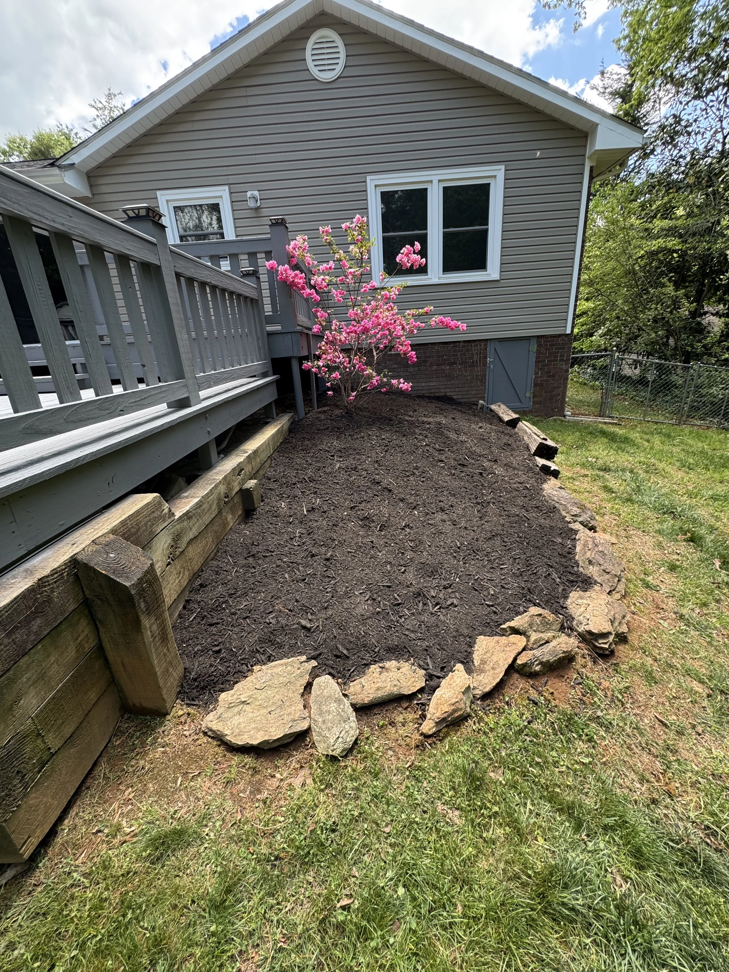 A backyard with freshly tilled dirt and a flower bed with pink flowering bushes. There is a wooden deck with gray railings attached to a house with beige siding and white trim, and a small gray door at ground level.