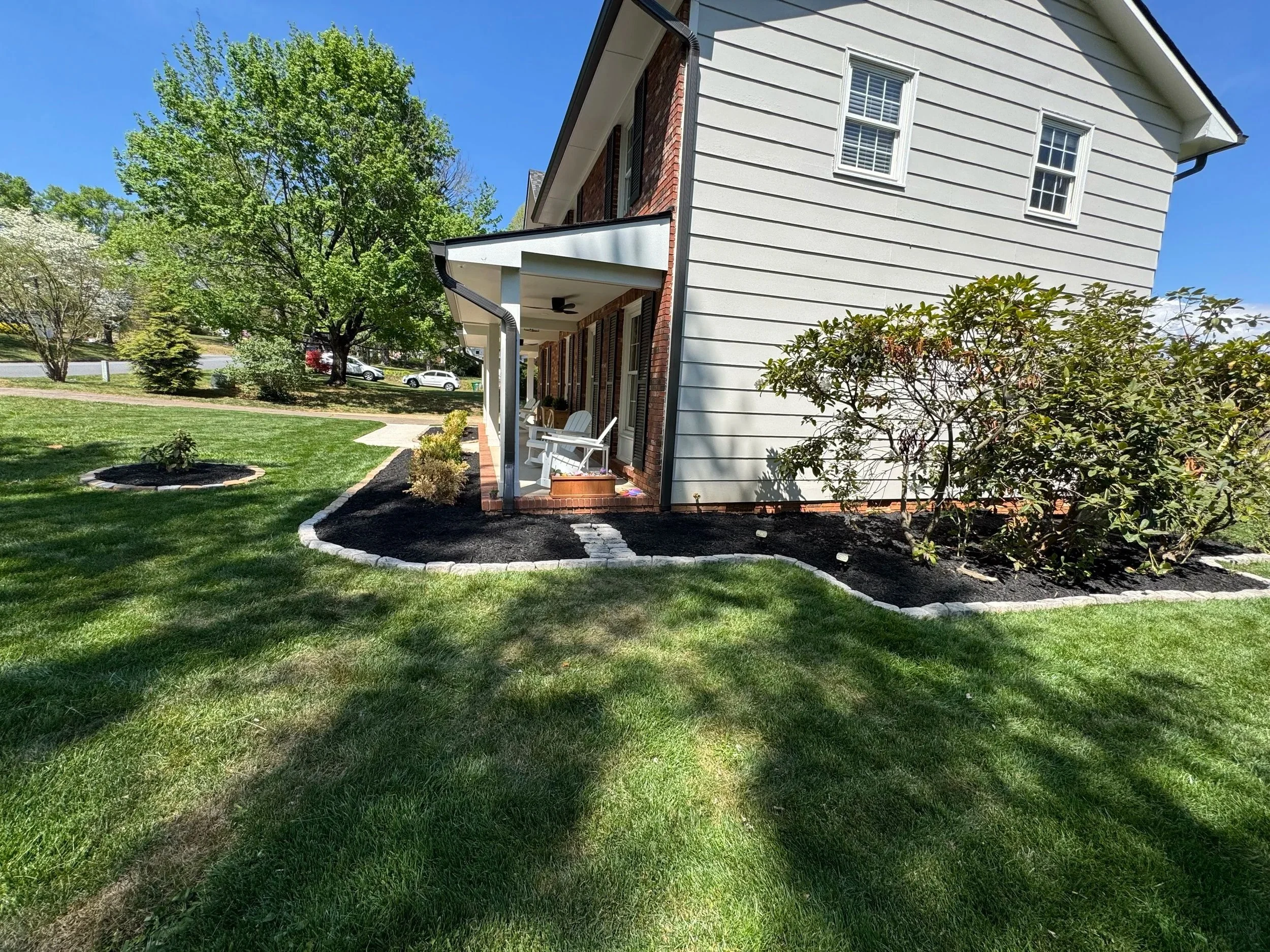 View of a house with white siding, red brick accents, and black shutters. Front porch with seating and landscaping. Large green trees and a well-maintained lawn under a clear blue sky.
