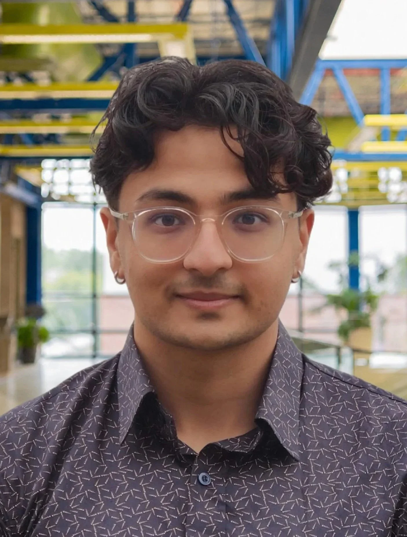 Close-up of a young man with curly dark hair, wearing glasses and a patterned dark shirt, smiling gently, in an indoor space with colorful metal framework in the background.