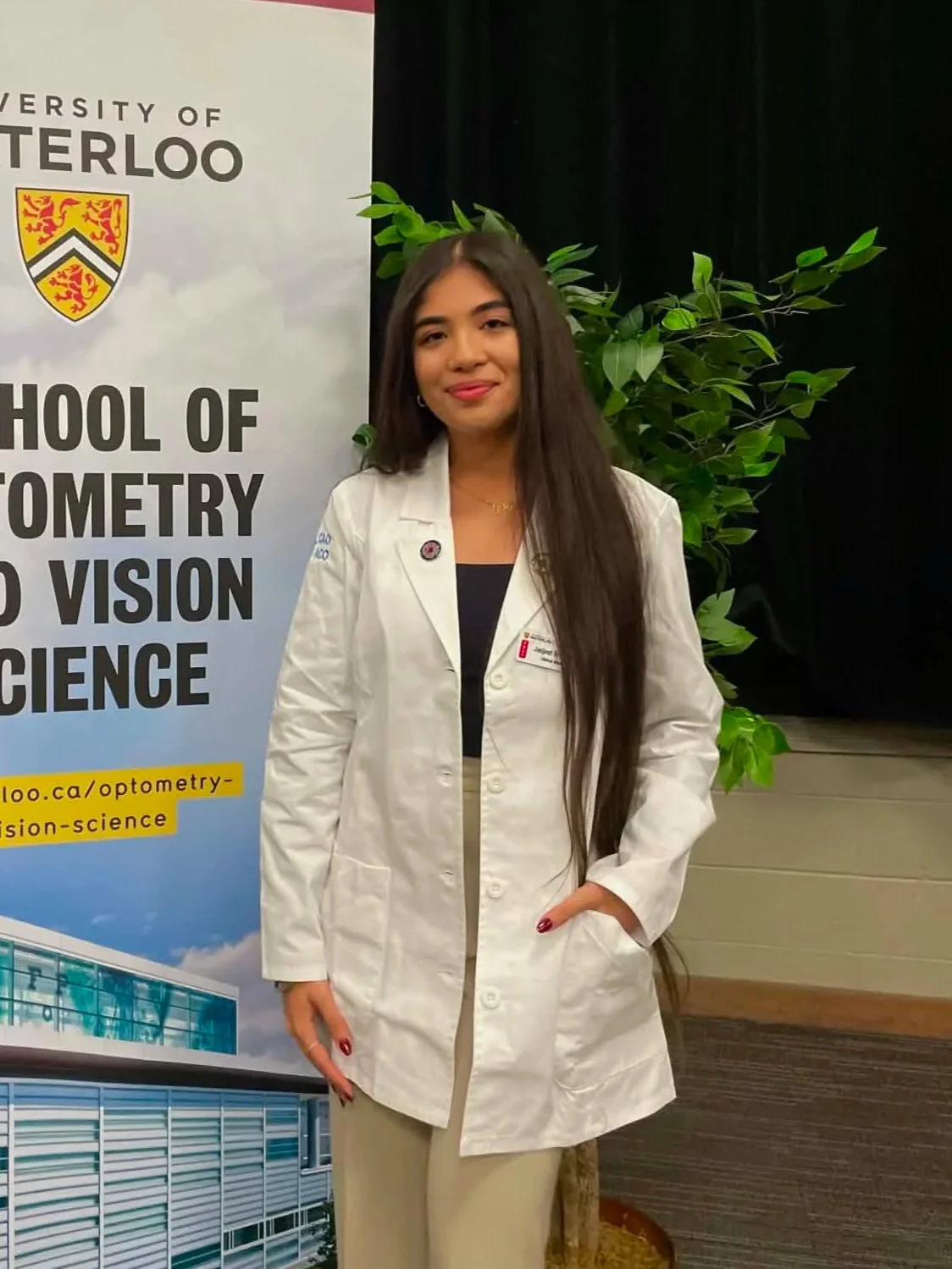 Young woman with long dark hair in white lab coat standing indoors next to a banner for the University of Waterloo School of Optometry and Vision Science with green plant in background.