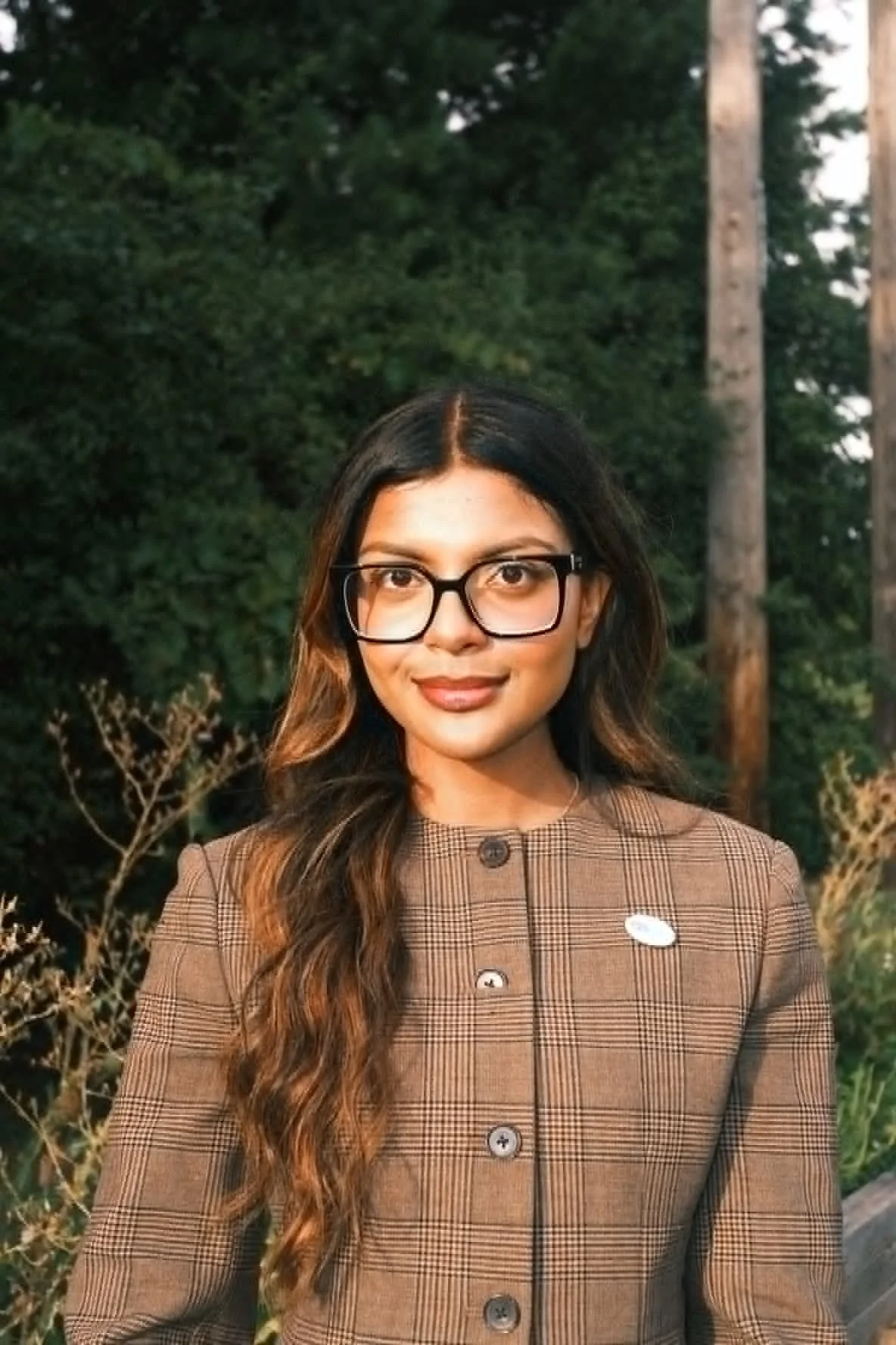 A woman with long wavy hair, wearing glasses and a plaid brown jacket, standing outdoors with trees in the background.