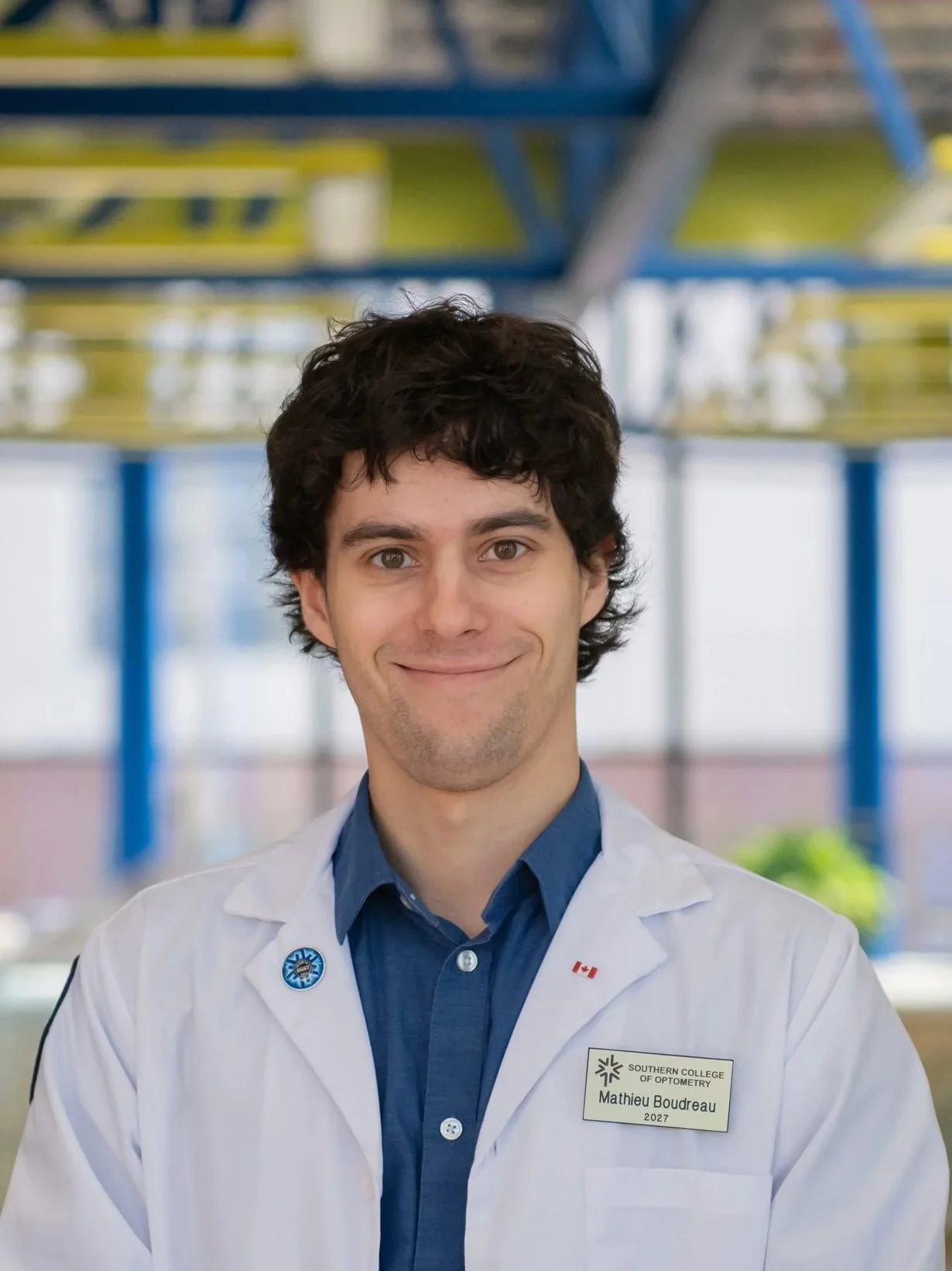A young man with dark, curly hair wearing a white lab coat with a name tag that reads Mathieu Boudreau, a blue shirt, and a pin that says Southern College of Optometry, standing in an indoor setting with blurred railings and structures in the background, smiling at the camera.