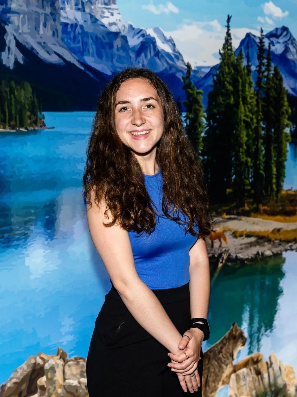 A young woman with long curly brown hair smiling in front of a scenic mountain landscape with a lake, tall pine trees, and snow-capped mountains.