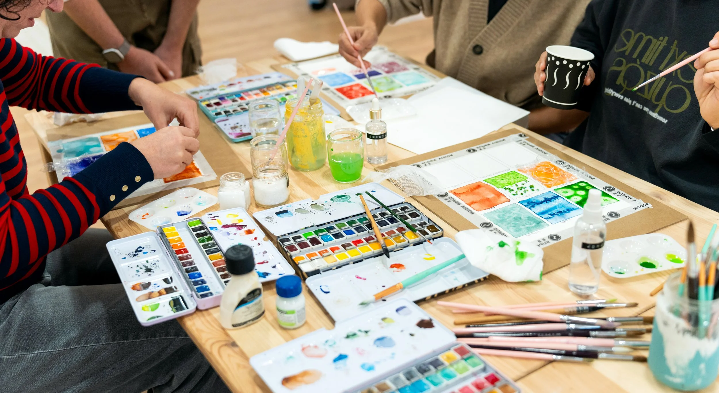 People participating in a watercolor painting activity at a table filled with various watercolor paints, brushes, and art supplies.