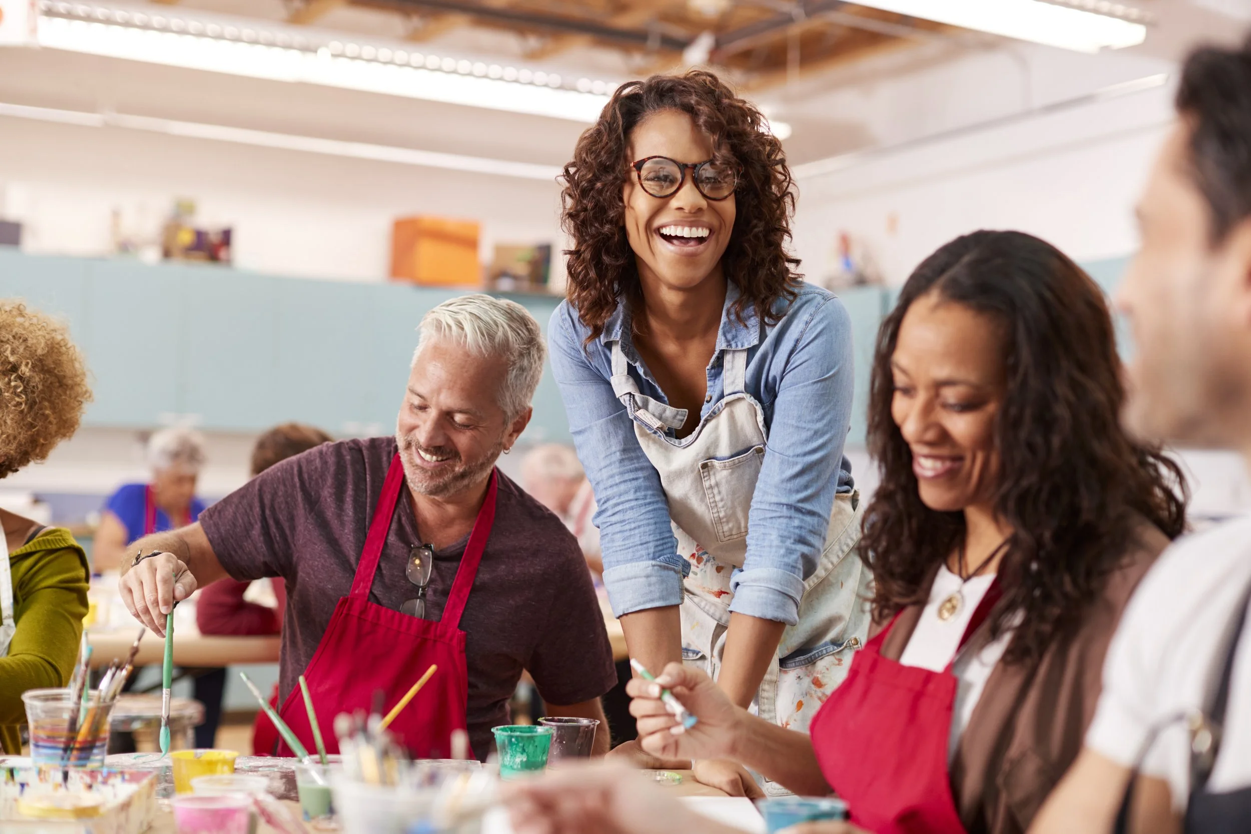 A group of diverse people, including women and men, enjoying a painting activity in a bright, cheerful art studio. They are using brushes and paint cups, with an instructor guiding them and smiling.