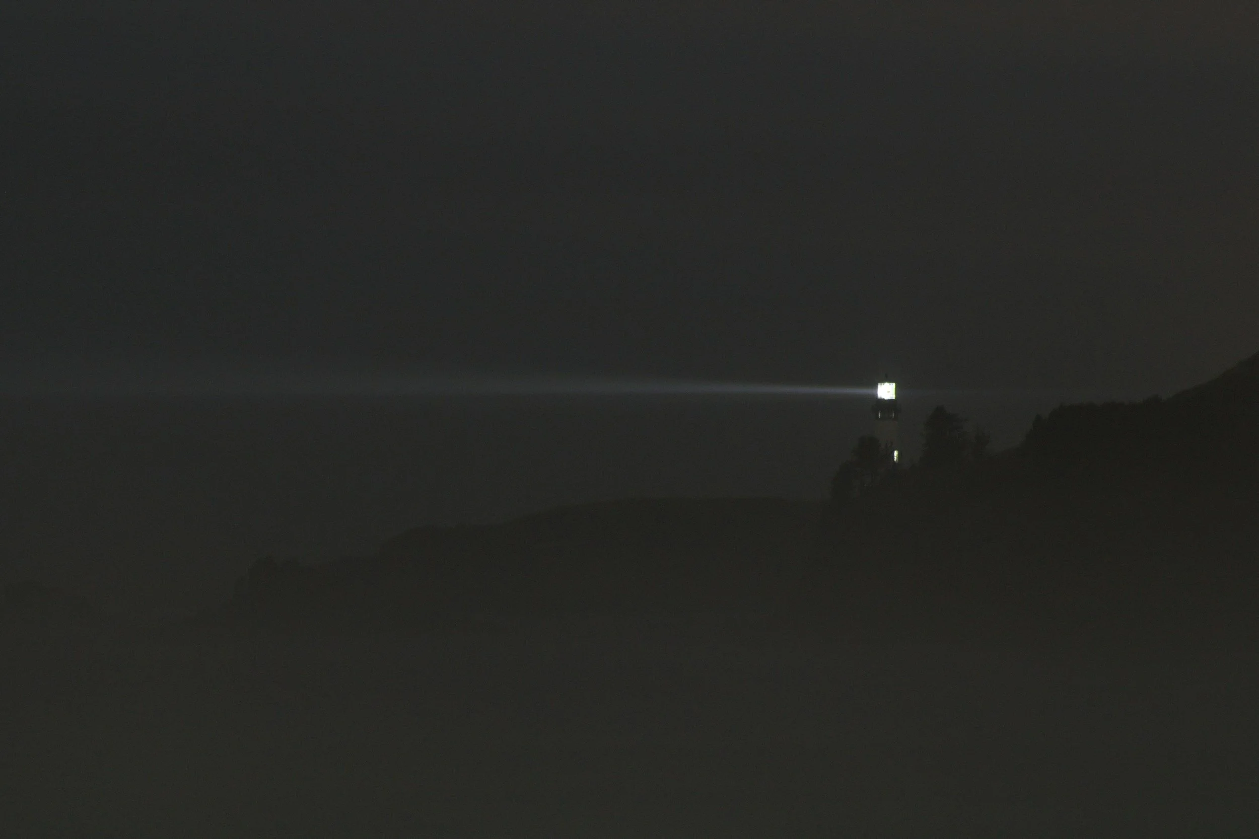 Nighttime scene of a shoreline with a lighthouse emitting a bright beam of light across the dark water, surrounded by silhouetted trees and hills.