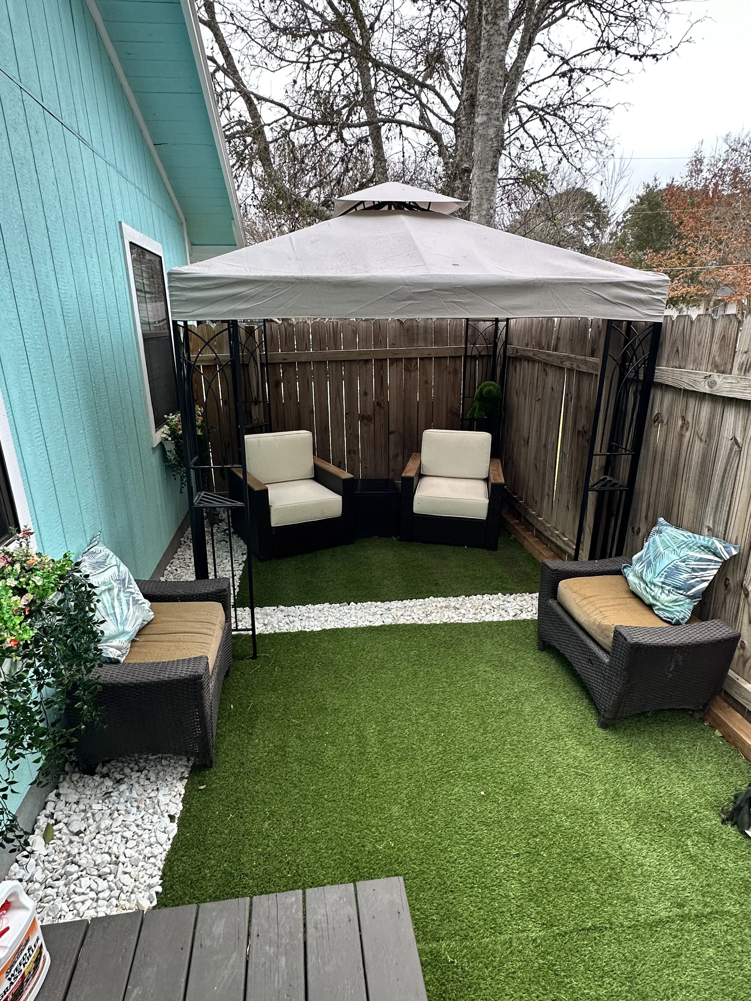 A backyard patio area with a blue house wall on the left, a wooden fence on the right, and a gravel border with white rocks. There is a green artificial grass area with two chairs in the foreground, and a covered gazebo with two more chairs inside. A