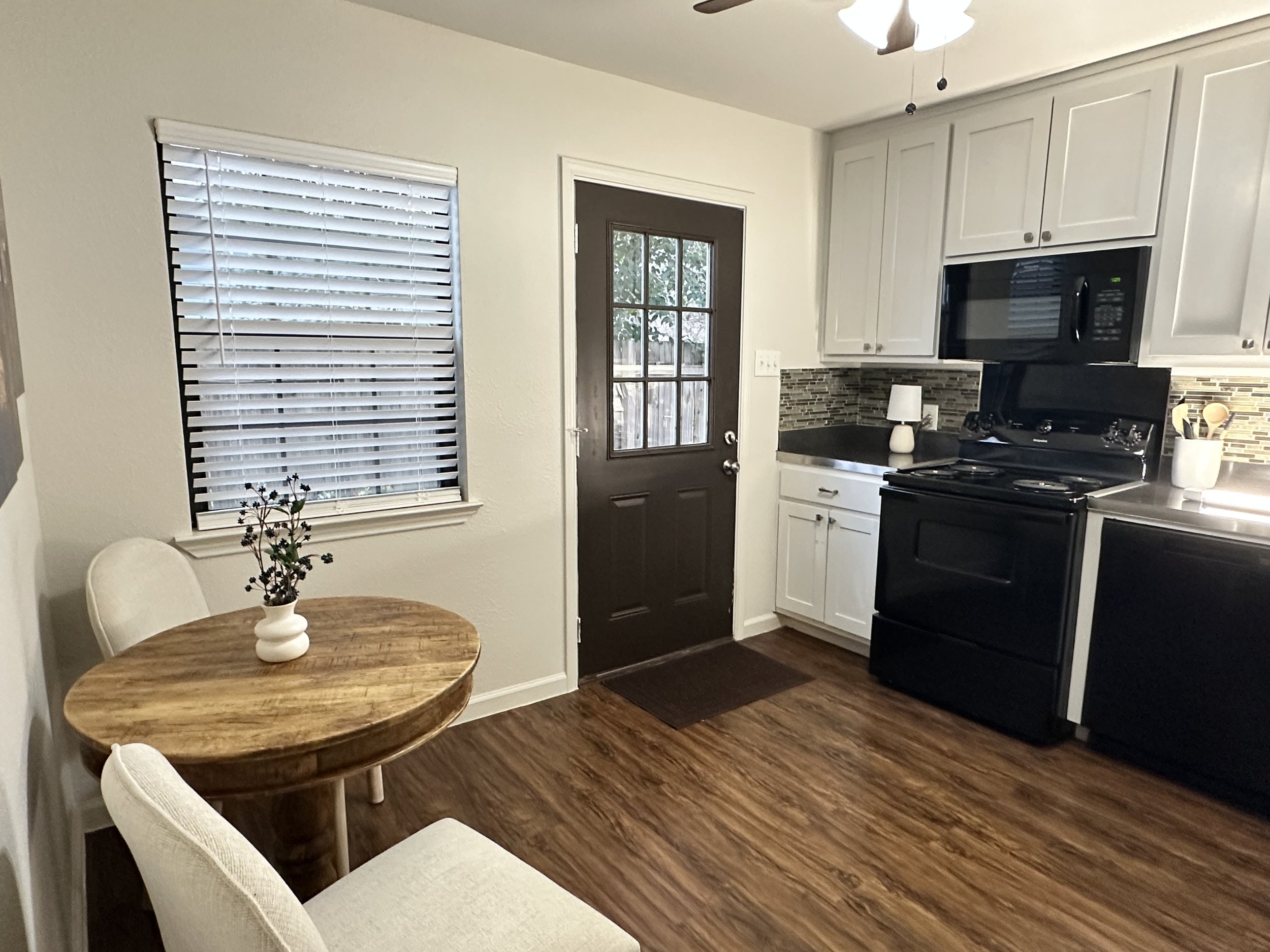 A small dining area and kitchen with wooden flooring, a round wooden table with a white vase and flowers, two white chairs, a window with white blinds, a dark door with a glass window, white cabinets, a black microwave, a stovetop, and a small white 