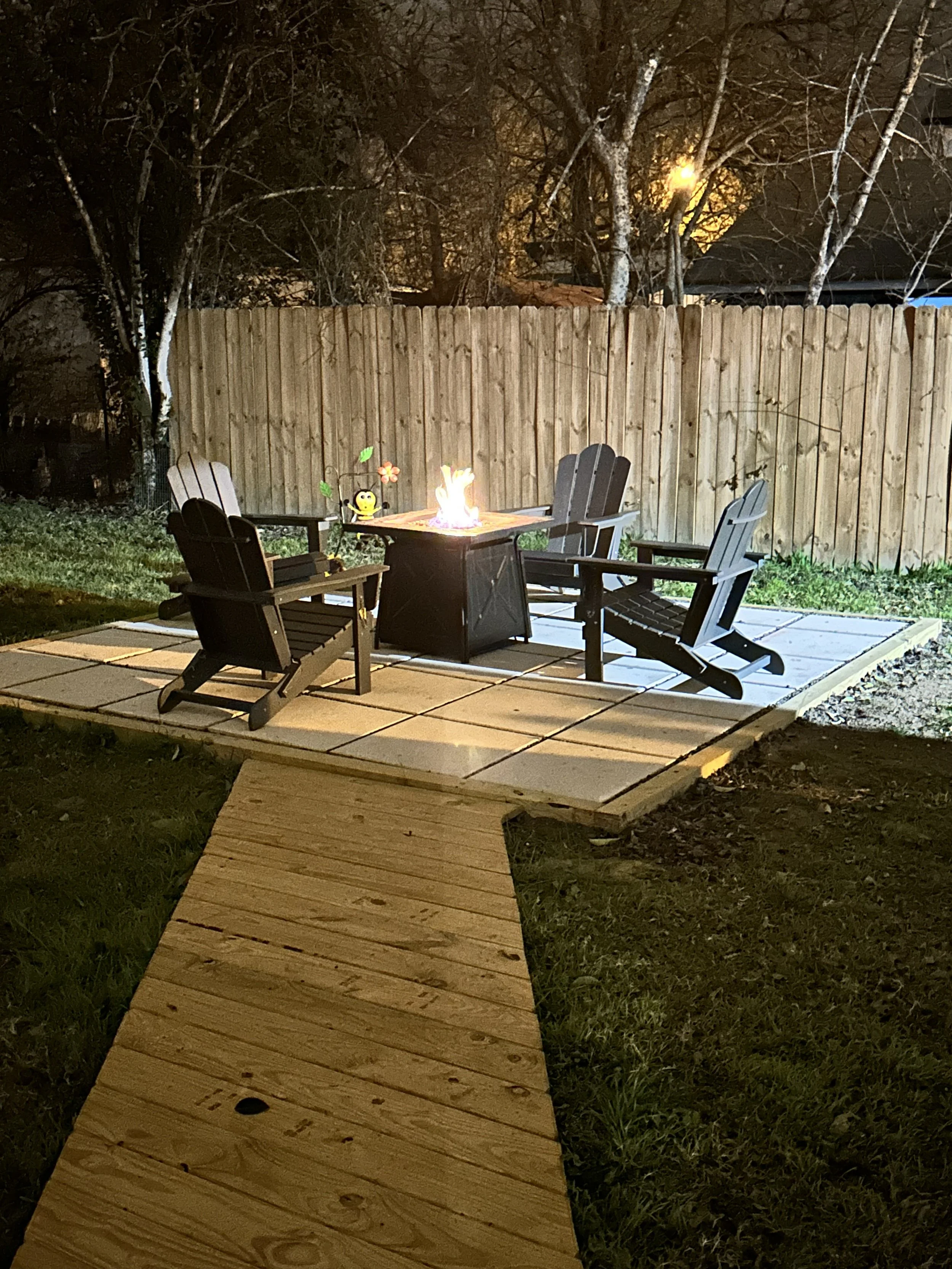 Nighttime backyard scene featuring a wooden walkway leading to a fire pit with chairs around it, a wooden privacy fence, trees, and a bright tree in the background illuminated by a streetlamp.