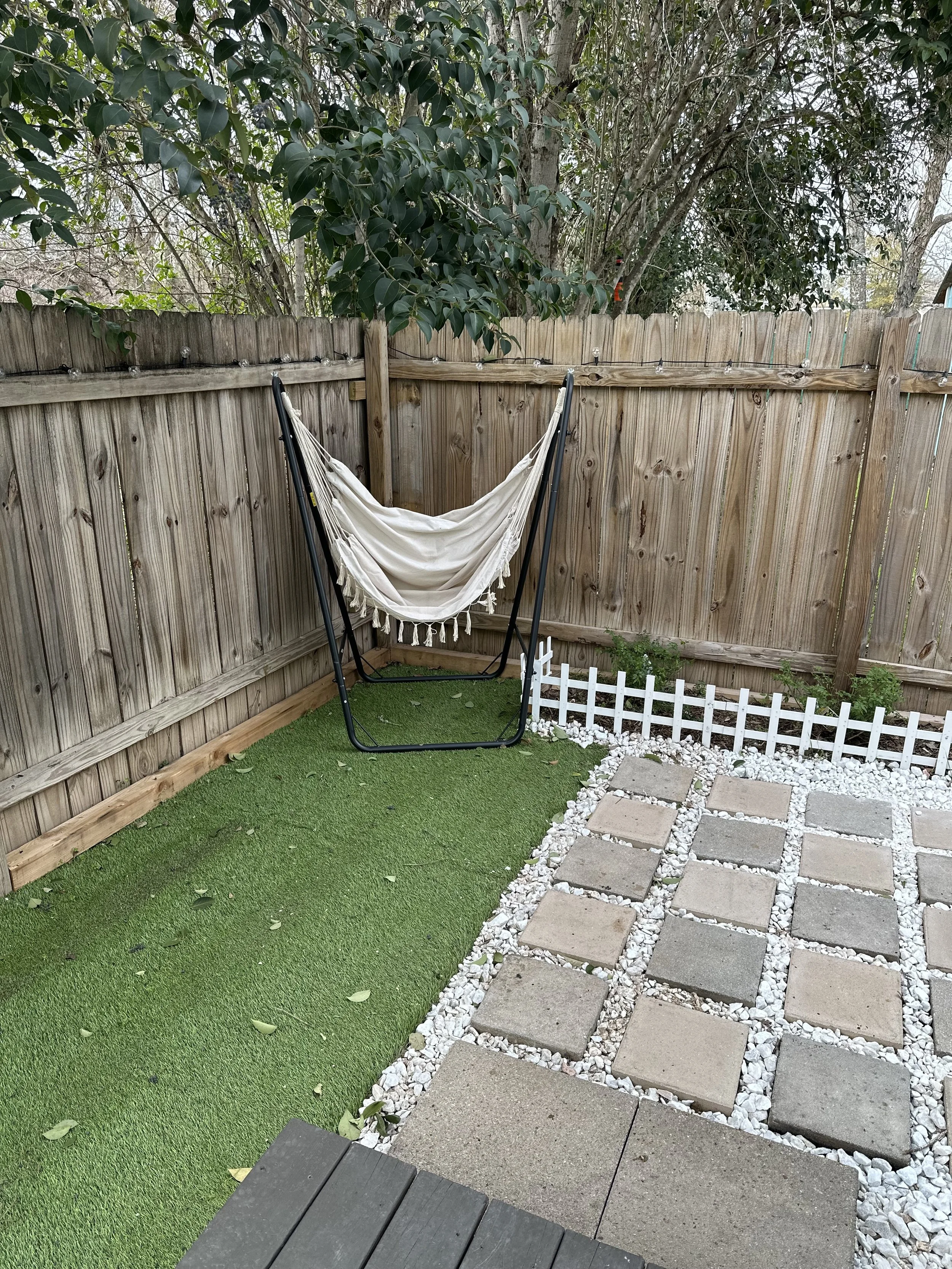 Backyard patio area with wooden fence, green artificial grass, a hammock, stone pathway, and small plants.