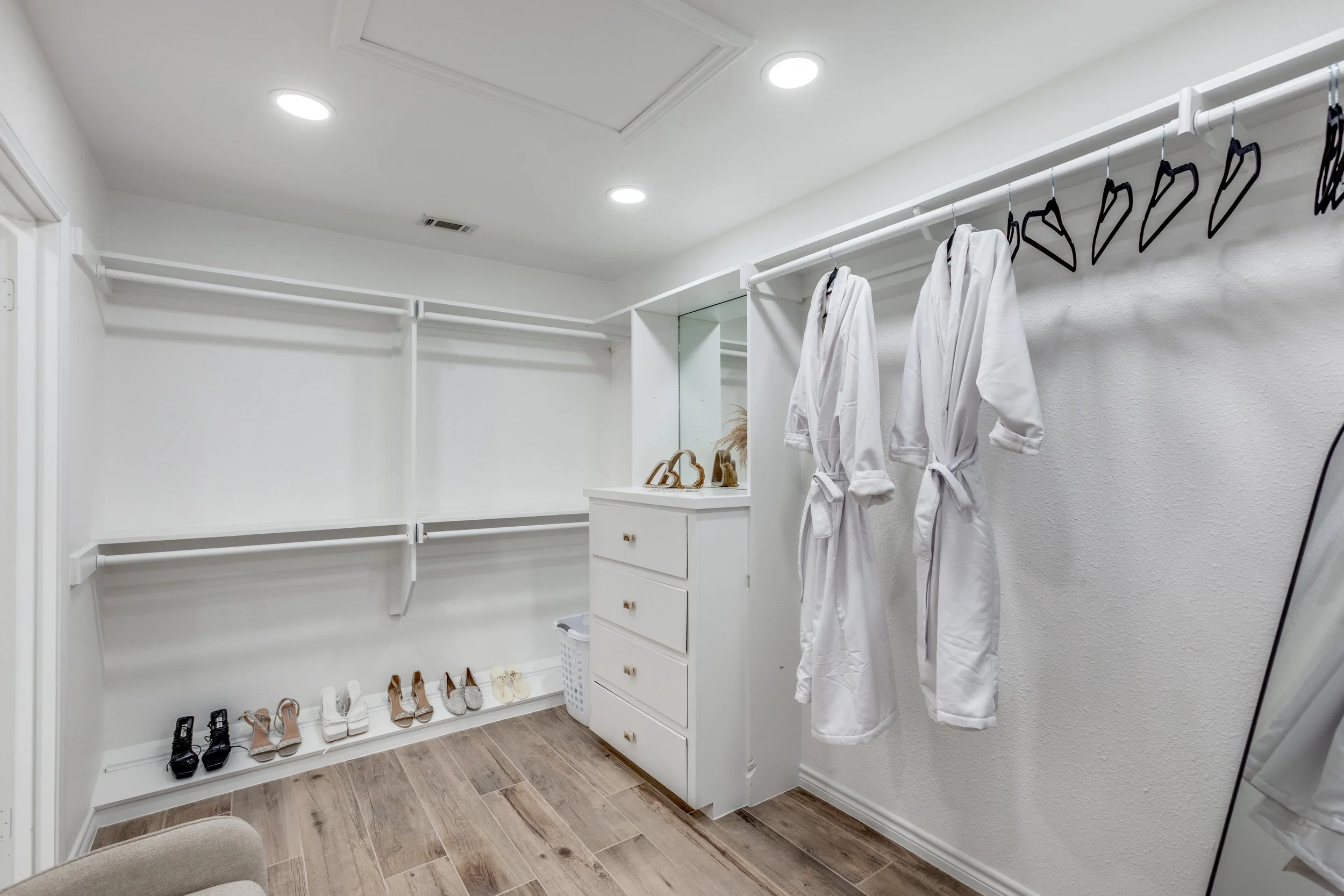 A walk-in closet with white shelves, hanging rods, and a small white drawer unit. Two white robes hang on black hangers, and shoes are arranged on the floor under the shelves, including high heels and sandals. The room has wood-like flooring and a mi