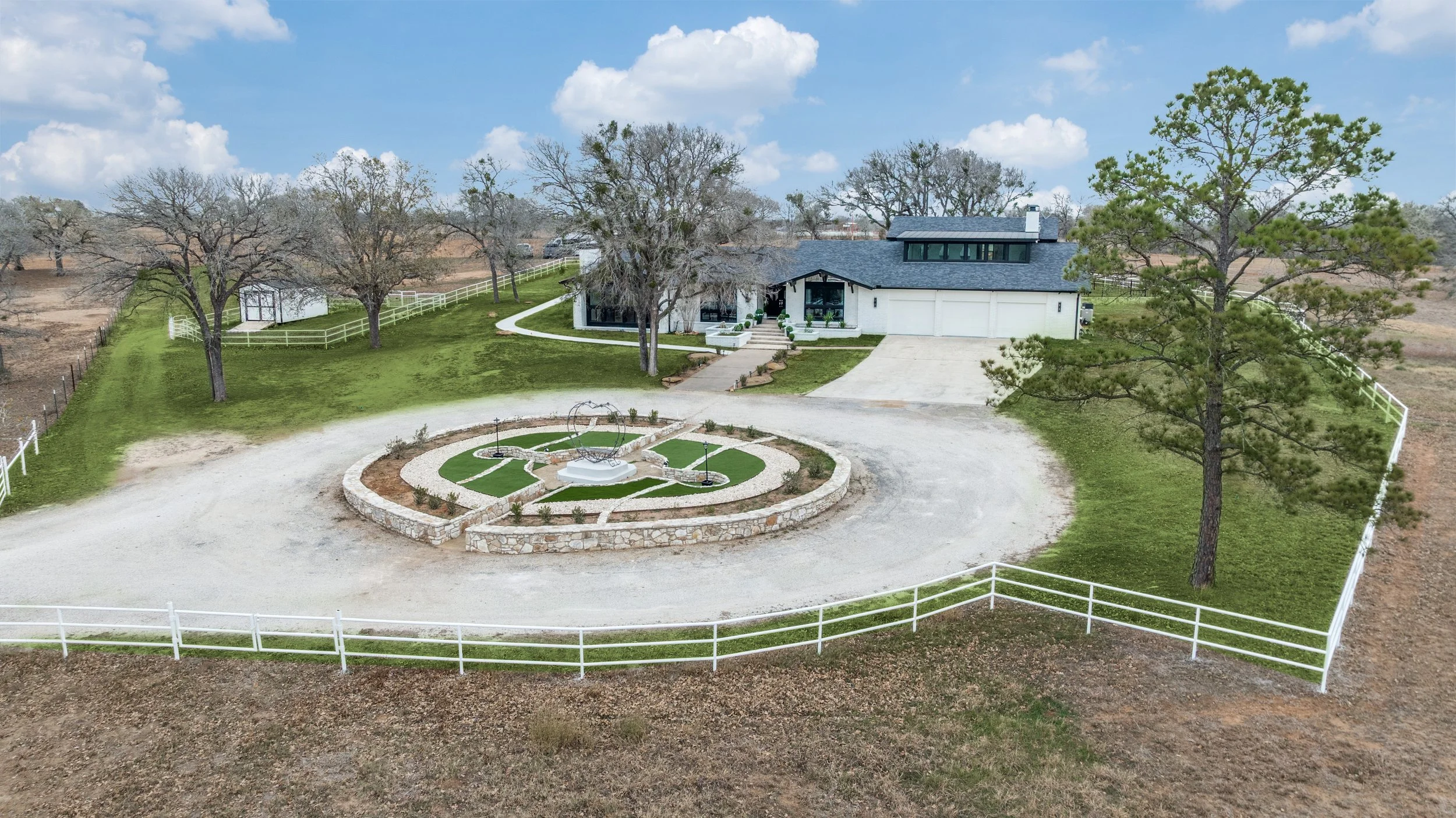 A spacious backyard with a white house, large trees, a circular gravel driveway, and a decorative garden bed with a sundial and benches.