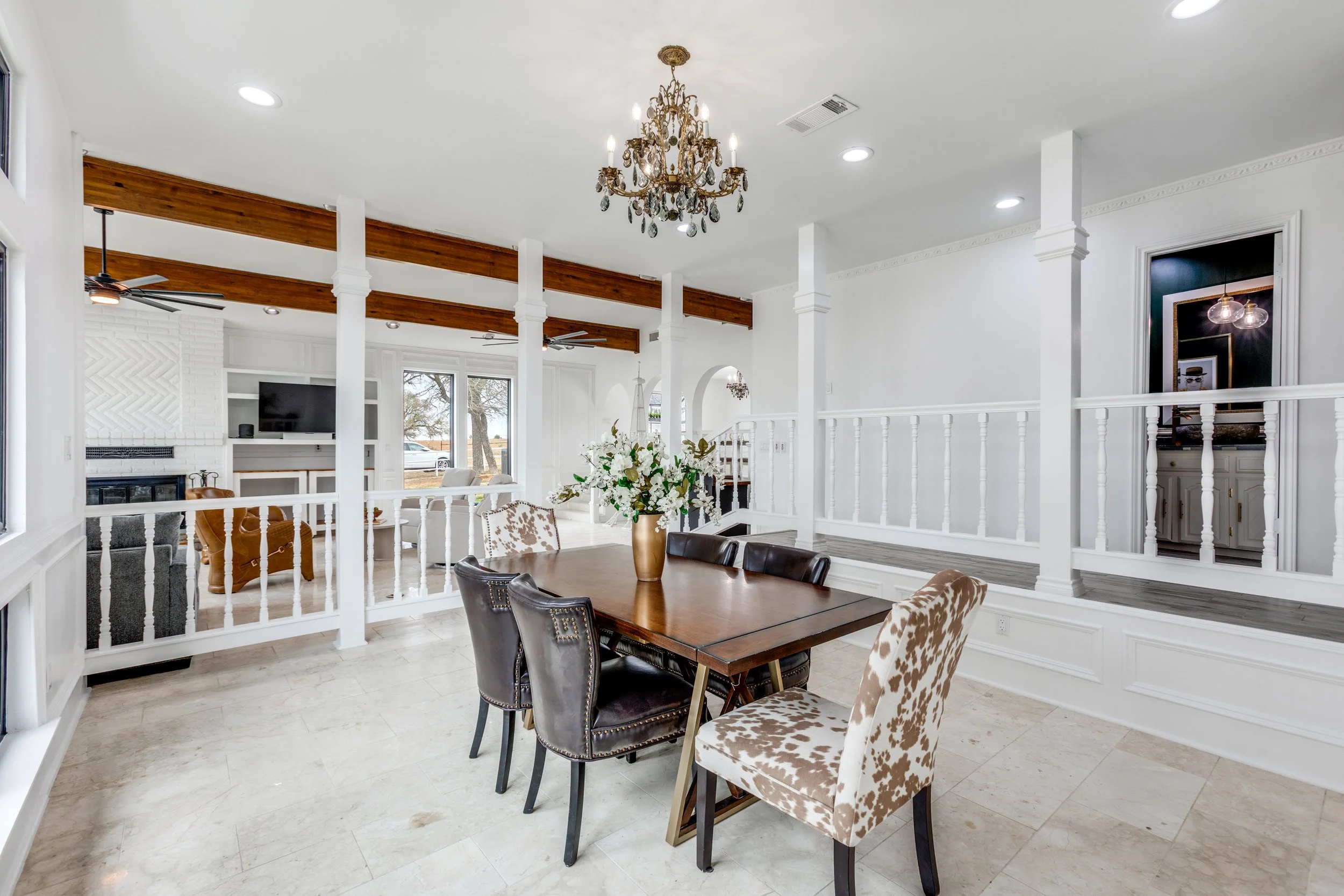 Bright, open living and dining area with white walls, beamed wooden ceiling accents, a chandelier, a fireplace, a flat-screen TV, and a large window showing trees outside.